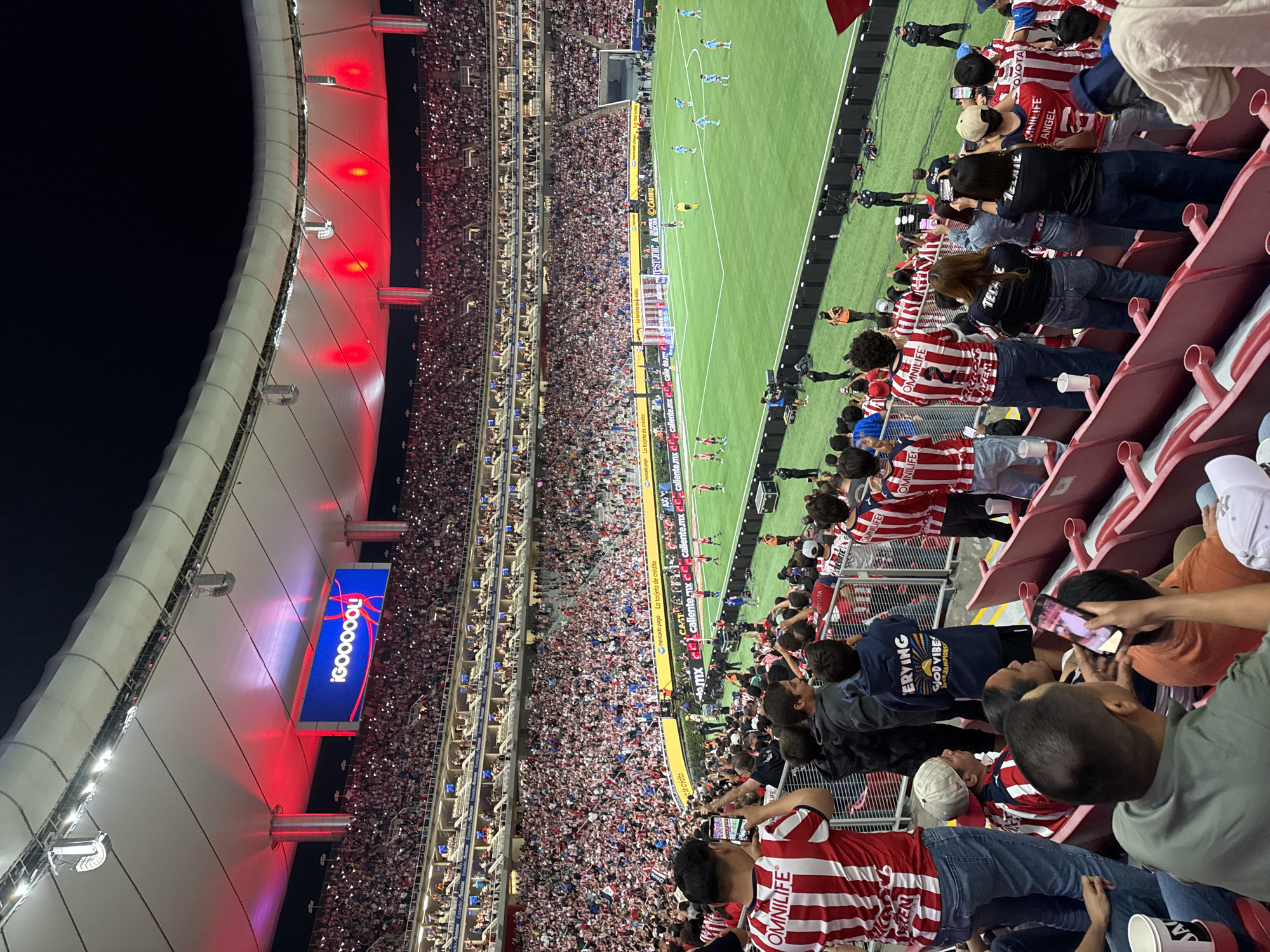 This is a high-angle view inside a stadium, likely during a soccer game, located in Zapopan, Mexico, as evidenced by the advertisements in Spanish. The stadium is densely packed with spectators, many wearing red and white striped shirts, possibly indicating support for the same team. In the foreground, fans are seated in red stadium seats, some holding up their phones to capture the action. In the distance, the soccer field is visible, with players engaged in a match. A large video board displaying "!GOOOOOL!" hangs from the stadium roof, which is illuminated with red lights. Numerous advertisements are placed around the field. The sky is dark, indicating the game is being played at night.