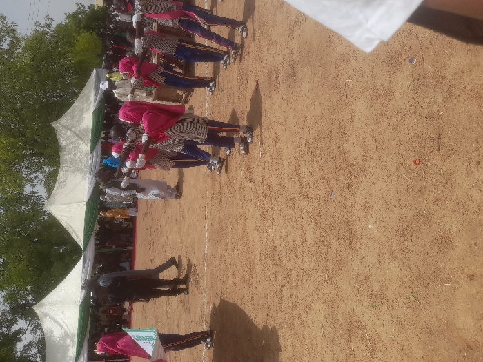 The image shows a group of young girls in uniform, standing in a line on a dirt field in Gusau, Nigeria (latitude: 12.187, longitude: 6.65). They are facing the camera and extending their arms forward, possibly holding sticks or flags. They are wearing black and white striped dresses with blue trim and pink head scarves. The girls are standing in front of a row of white tents, and there are adults and other people in the background. A man in a white uniform is standing with his arms crossed next to a girl holding a green flag in front of her. A man in dark clothing, likely security personnel, is watching them from a distance. The ground is dry and dusty.