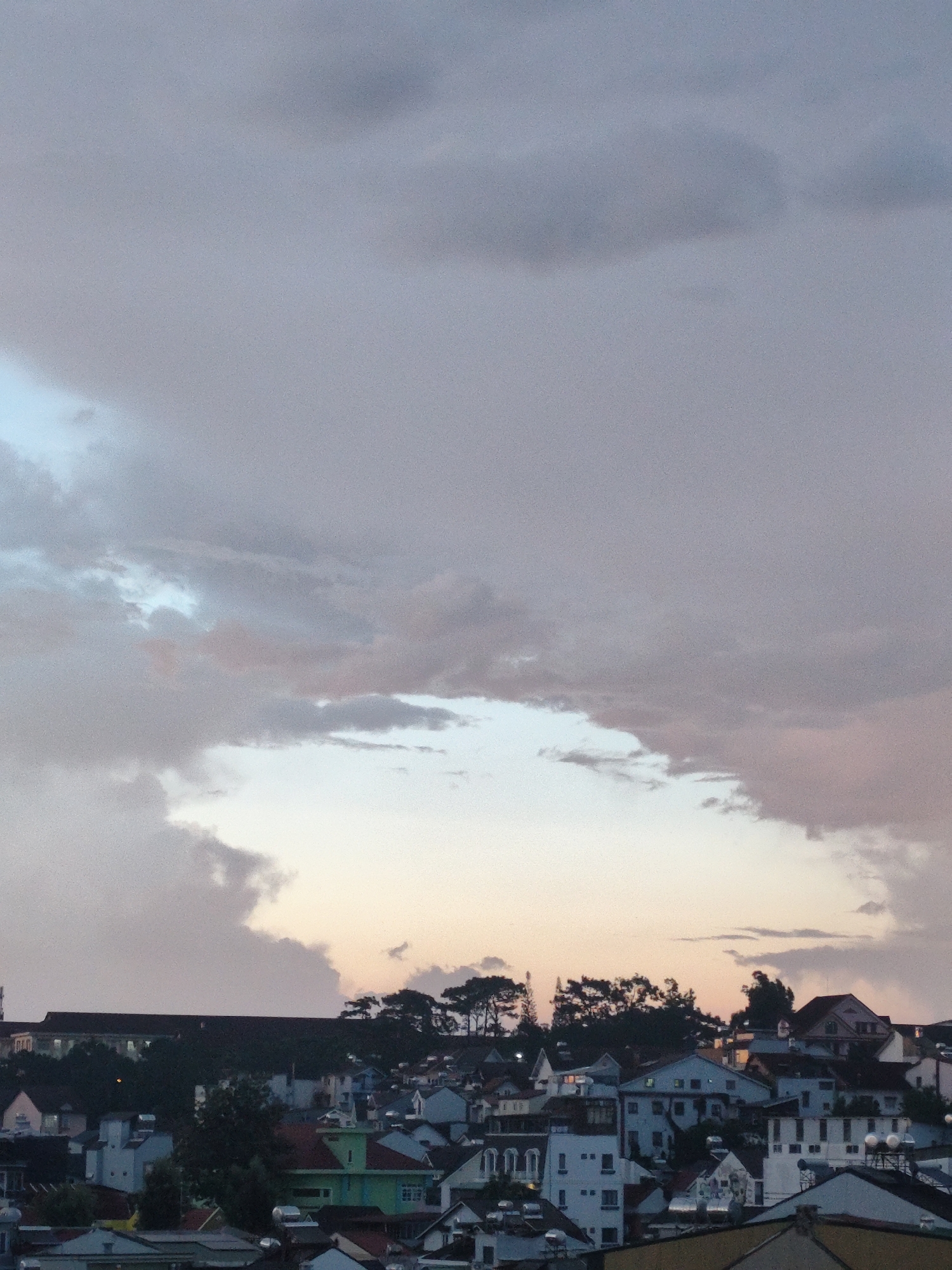 The image captures a twilight cityscape of Da Lat, Vietnam, marked by dense low-rise structures set against a backdrop of a predominantly cloudy, lavender-hued sky. The lower portion of the frame is filled with a collection of tightly packed houses, primarily white and gray in color, interspersed with occasional green and red roofs. In the distance, a dark, solid structure runs along the horizon line. Silhouette of trees is visible. The upper two-thirds of the image are dominated by gray and lavender clouds, creating a muted sunset or sunrise sky with a small patch of lighter, yellowish sky peeking through. The light suggests a time of day when daylight is waning or just beginning. There are no people visible in the shot. The overall impression is one of stillness and a landscape settling into or emerging from the quiet of night. The houses are densely packed with solar panels on many rooftops.