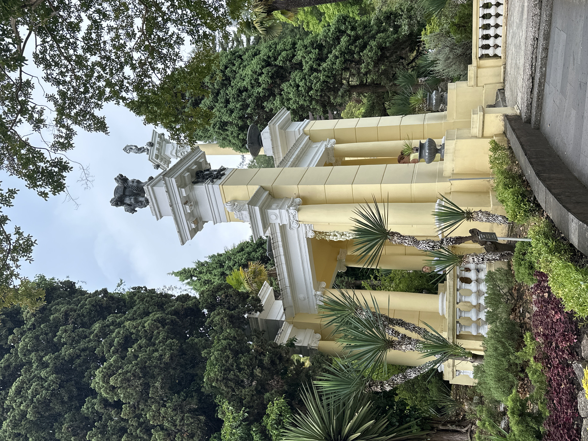 This image depicts a detailed view of the Monument to the Unknown Sailor in Sochi, Russia, an iconic structure located on the coast. The monument features a prominent yellow pavilion with classical architectural elements, including fluted columns and decorative white moldings. The pavilion is adorned with sculptures, notably a group of lions on one of the pedestals, and a standing female figure on another.
In the foreground, a lush garden with various plants, including spiky yucca-like trees and low-lying shrubs with red and green foliage, is visible. The plants create a verdant contrast with the bright yellow of the pavilion. Several grey urns are placed strategically around the structure, adding to its decorative appeal.
Behind the pavilion, dense greenery of tall coniferous trees forms a natural backdrop. The sky above is overcast, suggesting a cloudy day. A small section of a white balustrade is visible on the right side of the image, hinting at a pathway or terrace. A person can be seen partially obscured behind one of the pavilion's columns. The overall scene captures the architectural grandeur of the monument set within a beautifully landscaped environment.