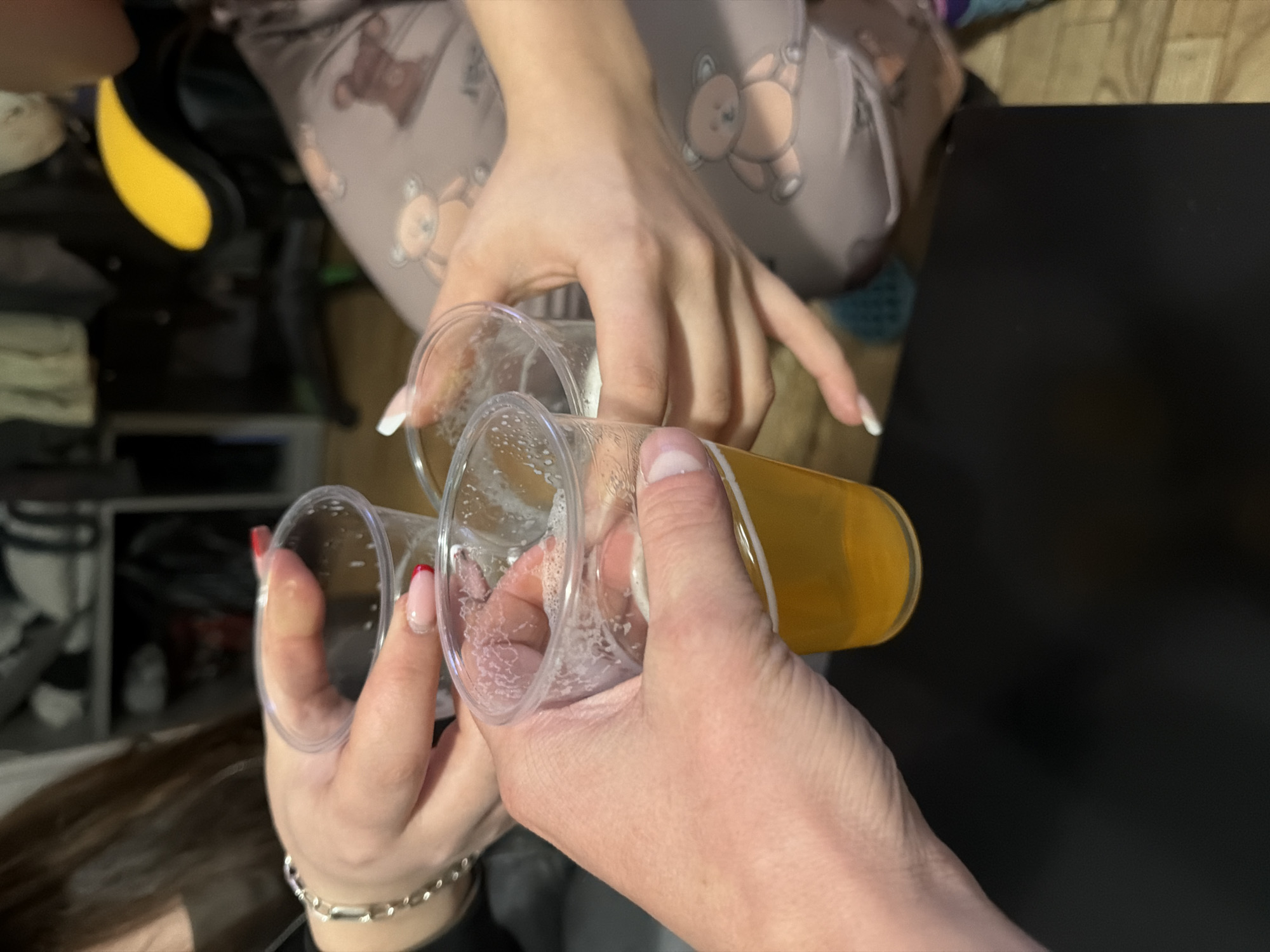 Three people are toasting with plastic cups filled with beer. Two people have manicured fingernails. One person has a silver bracelet. The environment includes a room interior with shelves, pajamas with teddy bear print, and a dark surface. It is not possible to discern from the image if the location is Varna, Bulgaria.