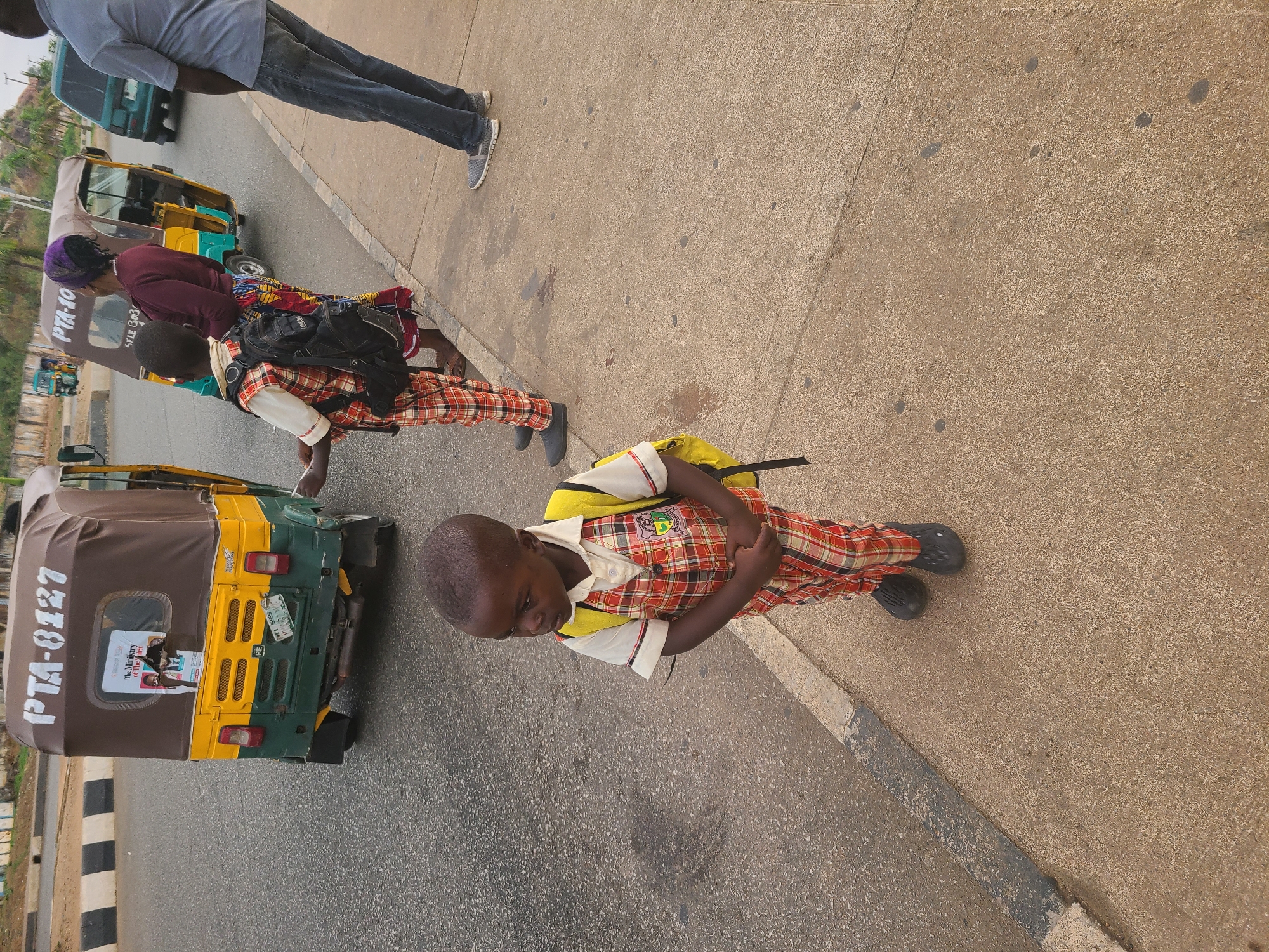 The image shows a boy in a red and white plaid school uniform standing on a sidewalk next to a street. He wears a yellow backpack and black shoes. An auto rickshaw, bearing the registration number "PTA-8127", is visible on the street next to the sidewalk. A woman wearing a purple head covering and carrying a black backpack stands further down the sidewalk. An additional man can be seen walking along the sidewalk. The road is asphalt and the sidewalk is concrete. The image was taken outdoors, during daylight hours. Based on the road features and auto rickshaw markings, the location is Jos, Nigeria.