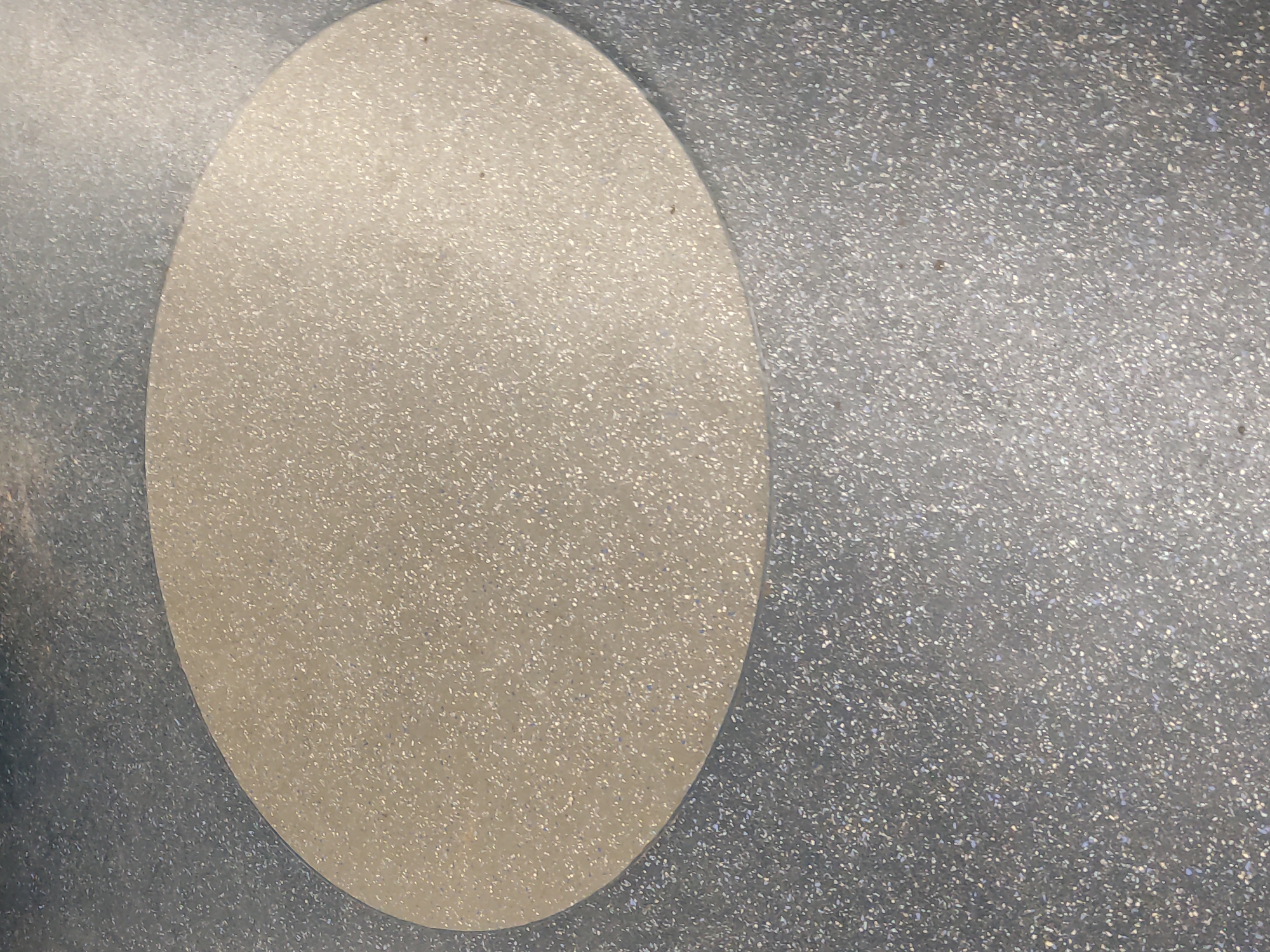 The image shows an oval-shaped, light-colored, speckled surface on a gray, speckled background. The texture appears to be a type of synthetic material or coating, with small reflective particles embedded throughout. The location is not evident from the image and I cannot provide the location in Moscow, Russia.