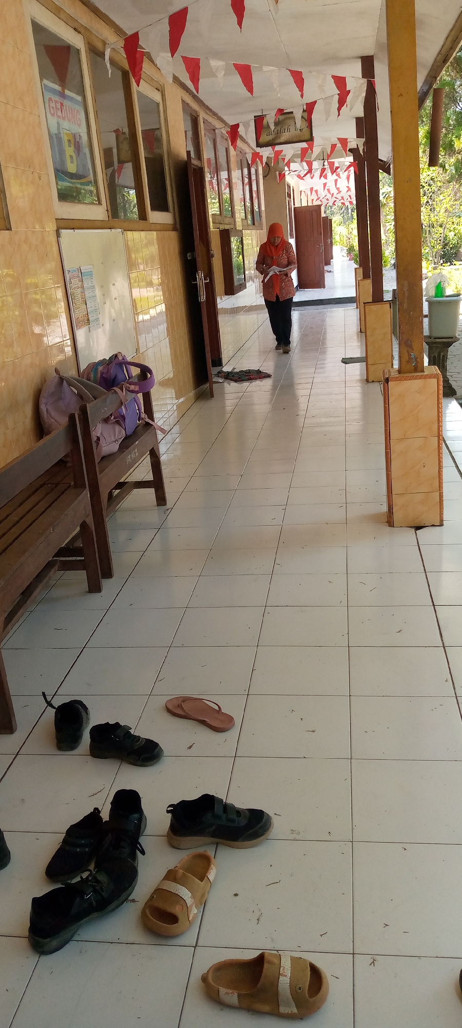 In Pacing, Indonesia, a woman wearing an orange headscarf and a patterned orange top walks down a tiled hallway. She is holding a white paper. The hallway has a tiled floor and walls with a yellowish hue. Red and white triangular flags are strung across the ceiling. On the left side of the hallway, there is a wooden bench with two purple and pink bags on it. Several pairs of shoes and sandals are scattered on the floor, including black sneakers, brown flip-flops, and tan sandals. The hallway appears to be part of a school or public building.