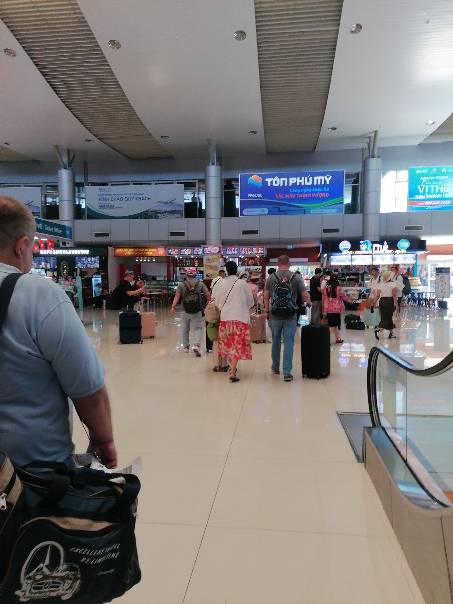 The image depicts the interior of Cam Ranh International Airport in Bac Cam Ranh Ward, Vietnam. Passengers with luggage are walking through the terminal. Several food and beverage vendors are visible in the background, along with signage in Vietnamese. An escalator is on the right side of the image. A man with a shoulder bag that has a Mercedes-Benz logo is on the left of the image.
