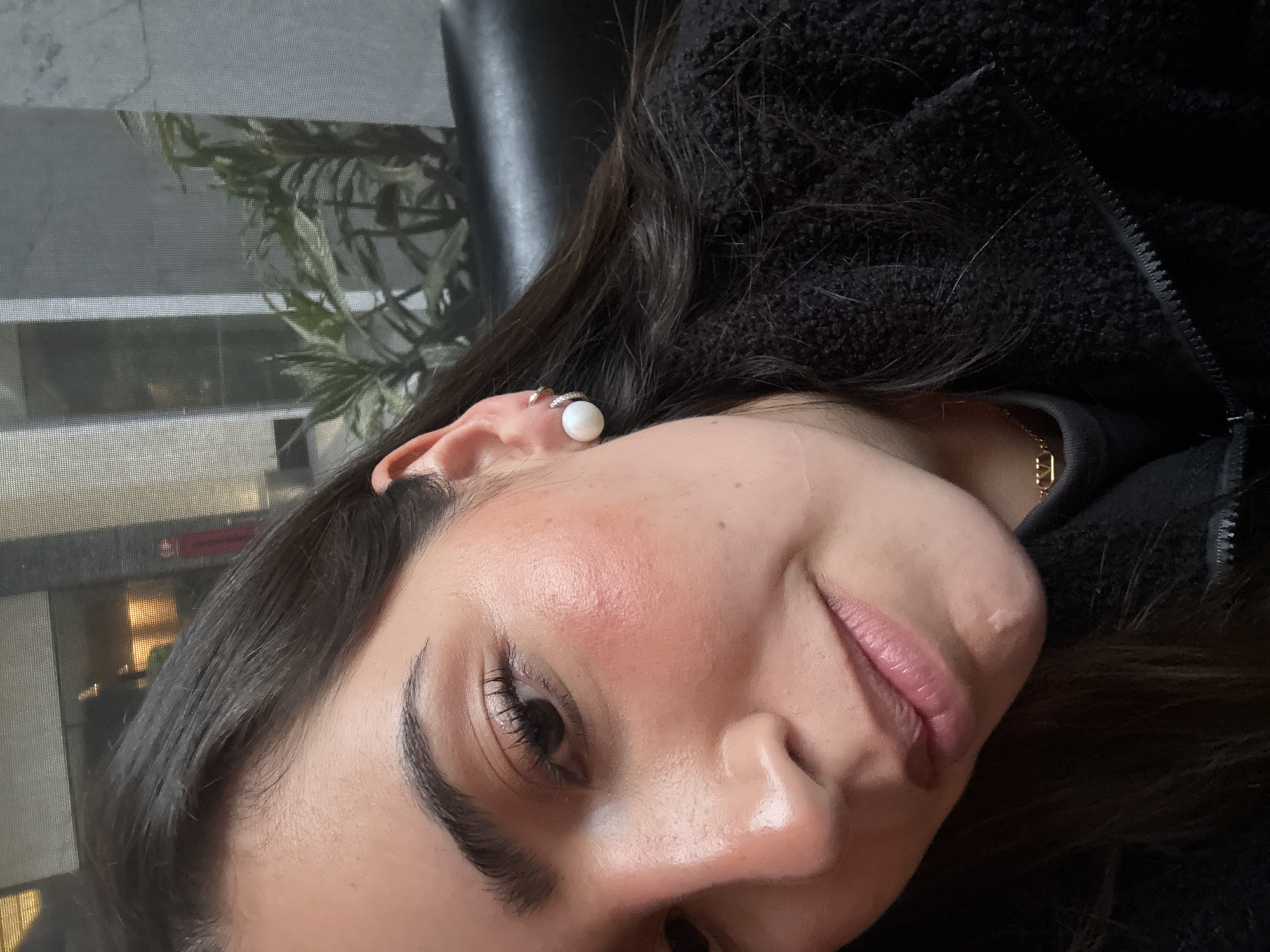 A close-up, slightly tilted portrait shot of a woman with dark hair and light skin. She is wearing a black fuzzy jacket and has a pearl earring on her left ear. There's a small hoop earring above the pearl one. A gold chain necklace is visible at the base of her neck. The background is blurred, showing a window with plants visible through it and part of a black leather-like surface. There is no visual information in this image to confirm the location is in the city of Puebla, Mexico.