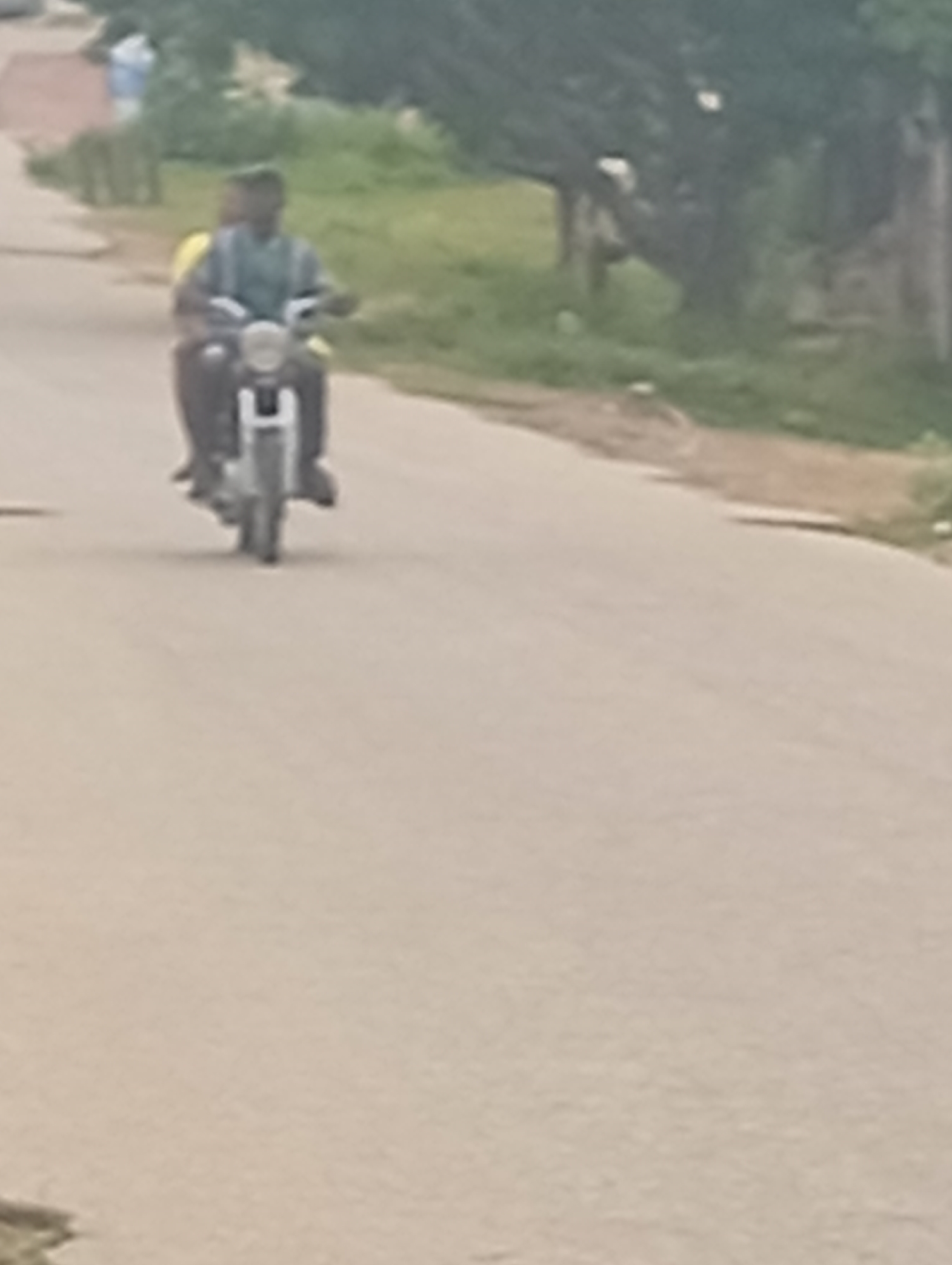 Two individuals, one driving and one as a passenger, are seen on a motorcycle on a paved road. The driver, who appears to be a male with dark skin, is wearing a blue and white striped shirt. The passenger, also with dark skin, is wearing a yellow top and is seated behind the driver. The motorcycle is silver with a black seat. The road is light gray and appears to be in a suburban or rural area. To the left of the road, there is a grassy area with some trees and structures that are out of focus. To the right of the road, there are more trees and a wall or building. The sky is overcast, and the overall lighting suggests it is daytime. The location is identified as Adankolo, Nigeria.