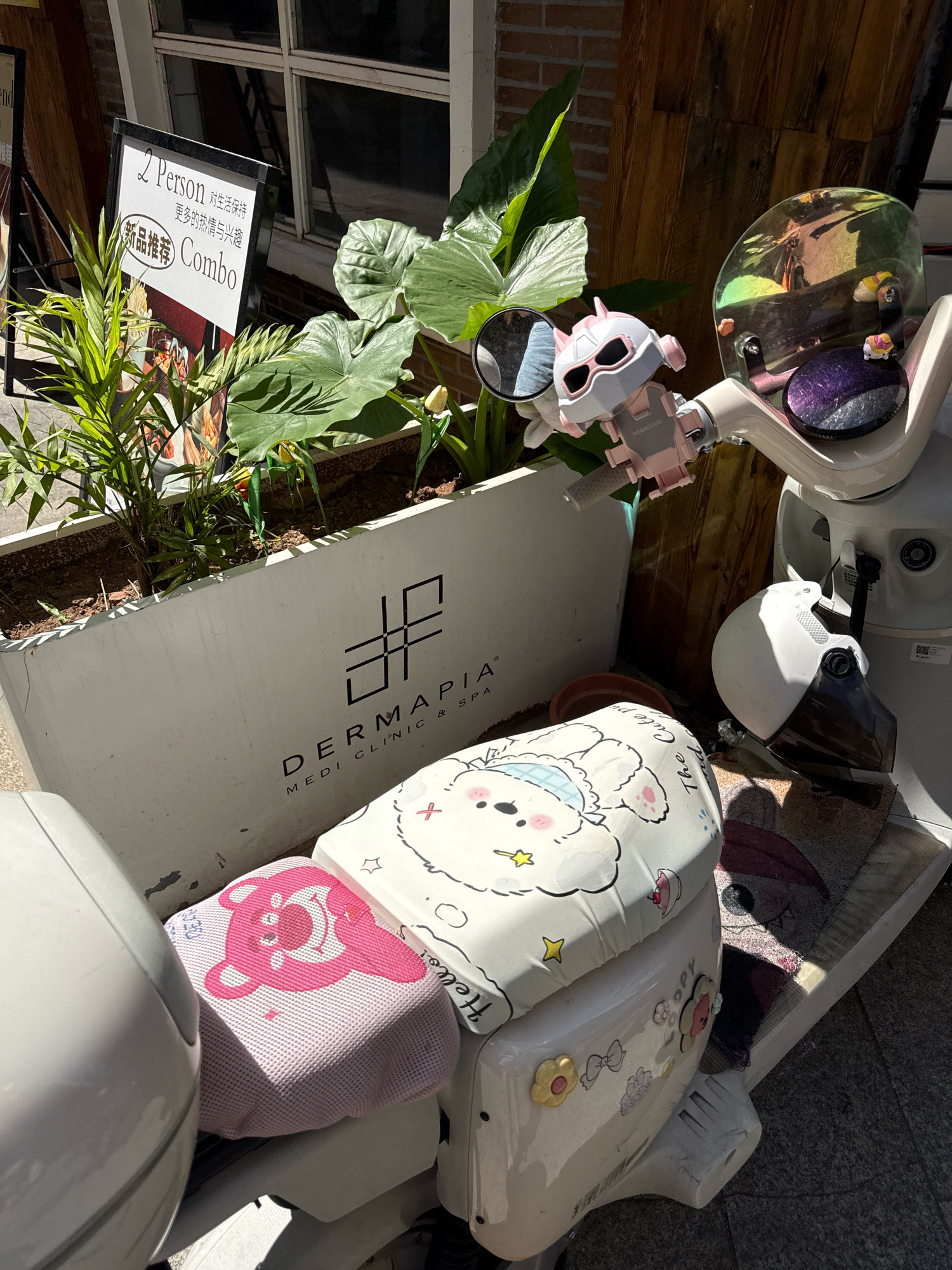 An outdoor scene shows a white electric scooter parked next to a white planter box. The scooter is decorated with a pink cartoon bear seat cover and a white cartoon bunny seat cover. A white helmet with a black visor is hanging on the side of the scooter. A toy pink robot is attached to the handlebars. The planter box contains green plants and has the logo "DERMAPIA MEDI CLINIC & SPA" on its side. A sign in the background advertises "2 Person Combo." The ground is paved with dark gray asphalt. The scene is lit by sunlight, creating shadows.
