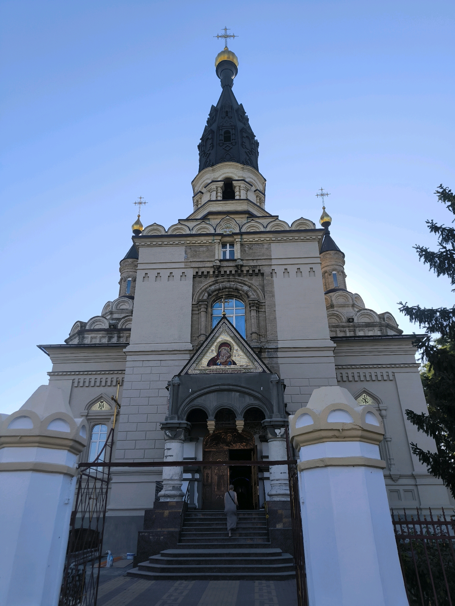 This is a low-angle, full shot of the Church of the Icon of the Mother of God "Joy of All Who Sorrow" located in Mykolaiv, Ukraine, on a clear, sunny day. The church is constructed from light-colored brick and stone, with multiple domes and a tall, dark, multi-tiered spire topped with a golden cross. A large mosaic of a religious figure is visible above the entrance. The entrance features ornate golden doors and is accessed by a wide set of stairs. In front of the church, there are decorative white and yellow pillars and a metal fence. A lone individual in a light-colored dress is walking up the stairs towards the church. The background consists of a clear blue sky with sunlight casting rays through a nearby evergreen tree.