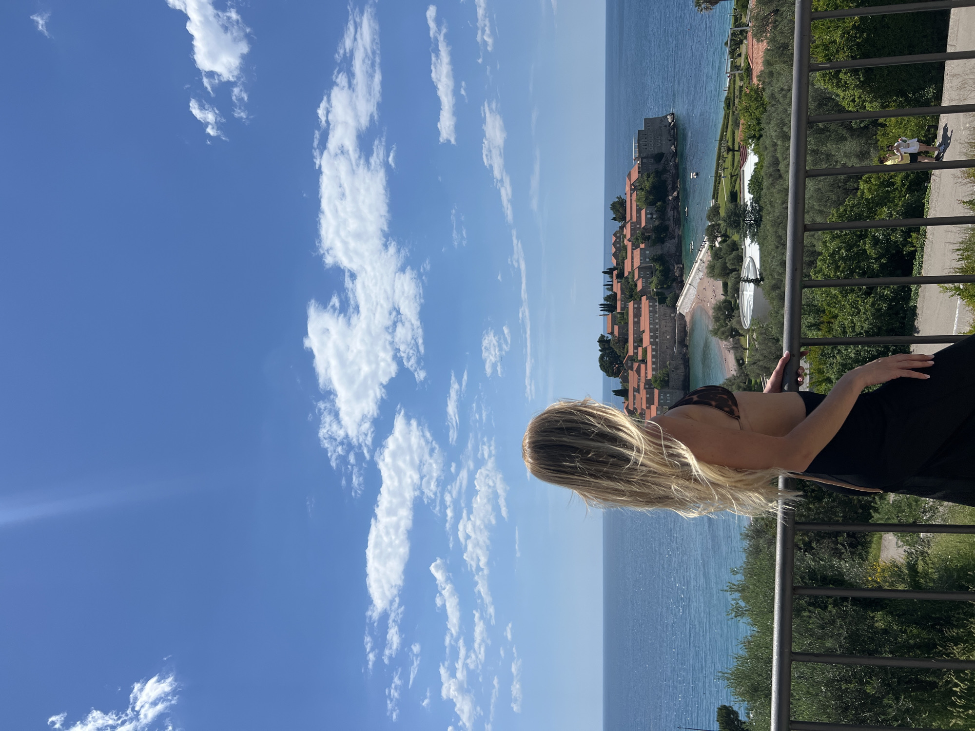 A woman with long blonde hair, wearing a bikini top and dark pants, leans on a metal railing overlooking Sveti Stefan, Montenegro. The sea stretches to the horizon under a bright blue sky with scattered clouds. The island of Sveti Stefan is visible, with its densely packed orange-roofed buildings and a beach. Two figures stand on a path behind the railing. The scene is filled with sunlight.