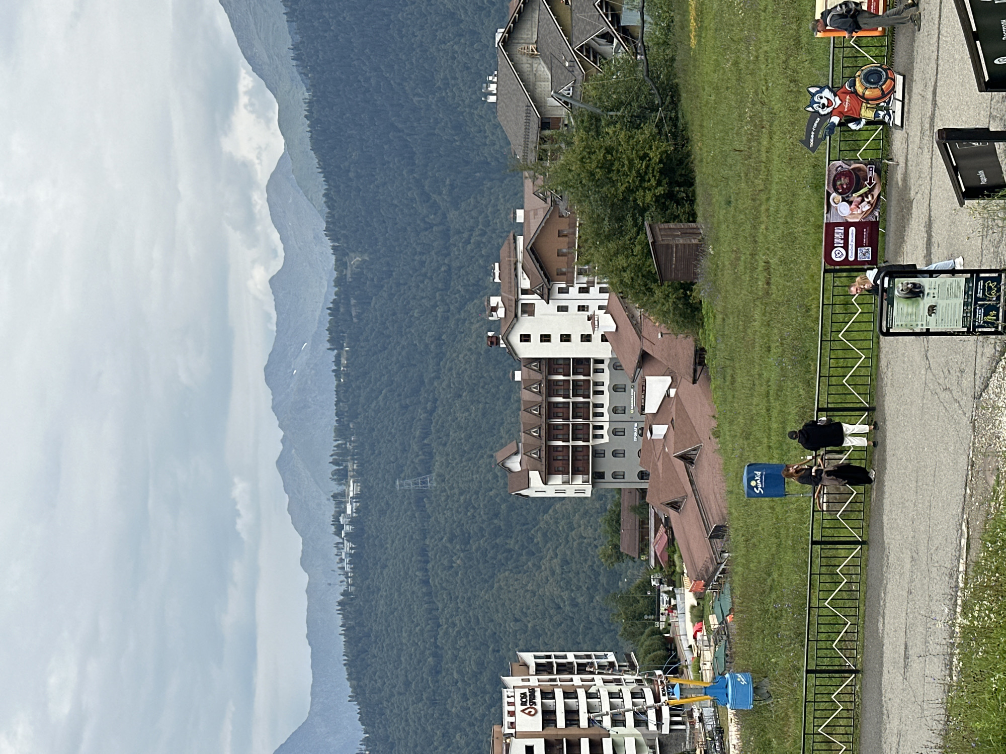 A vertical medium shot captures a mountain resort area under a cloudy sky. The foreground features a paved walkway and a grassy slope with a black metal fence. Several people are present, with two individuals standing near the fence, facing away from the camera. Further right, a cutout of a cartoon wolf mascot stands next to a sign. Signs are also visible on the walkway. In the midground, a cluster of buildings with red-tiled roofs and light-colored facades are nestled among green trees. Some of these buildings appear to be hotels or apartment complexes, with balconies and windows. A large, modern hotel building with a white facade and balconies is prominent on the left. A ski lift or gondola mechanism, painted blue and yellow, is visible near this building. The background is dominated by a dense, dark green forest covering a mountainside, with more distant, hazy mountain ranges under the overcast sky. A tall metal pylon is visible on the forested slope. The overall impression is of a resort town in a mountainous, possibly summer, setting.
