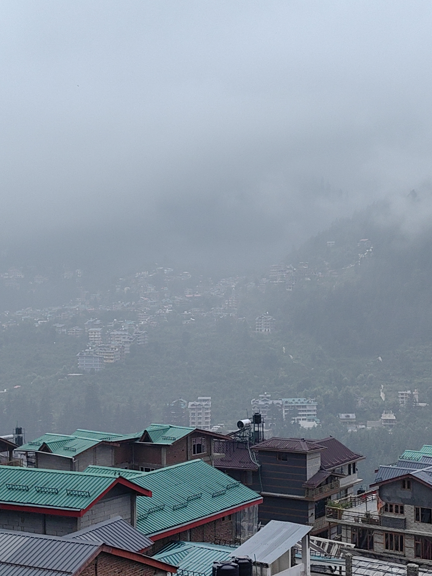 The image depicts a panoramic view of Vashisht, India, a town nestled in a mountainous region, characterized by its dense residential architecture and lush green slopes. The sky is overcast, with fog or mist partially obscuring the distant hills and buildings, creating a subdued and atmospheric scene.
In the foreground, several buildings with distinct roofing styles are visible. Many of these structures feature vibrant, teal-colored metal roofs, often with red trim along the edges. Some buildings display brickwork and grey concrete construction. One prominent building in the mid-foreground has a dark, multi-story facade with horizontal banding and a dark, pitched roof. Water tanks are visible on the rooftops of some of these foreground buildings.
Further into the background, the town extends up the hillsides. Numerous buildings, varying in size and design, are scattered across the slopes. These structures appear to be residential or hotels, with some featuring pitched roofs and balconies. The dense arrangement of buildings suggests a well-populated area. The natural landscape of forested hills provides a backdrop, though it is significantly softened by the atmospheric haze. A faint winding path or road can be seen ascending one of the hills. The overall impression is one of a bustling mountain town shrouded in the soft embrace of mist. There are no visible people in the image.