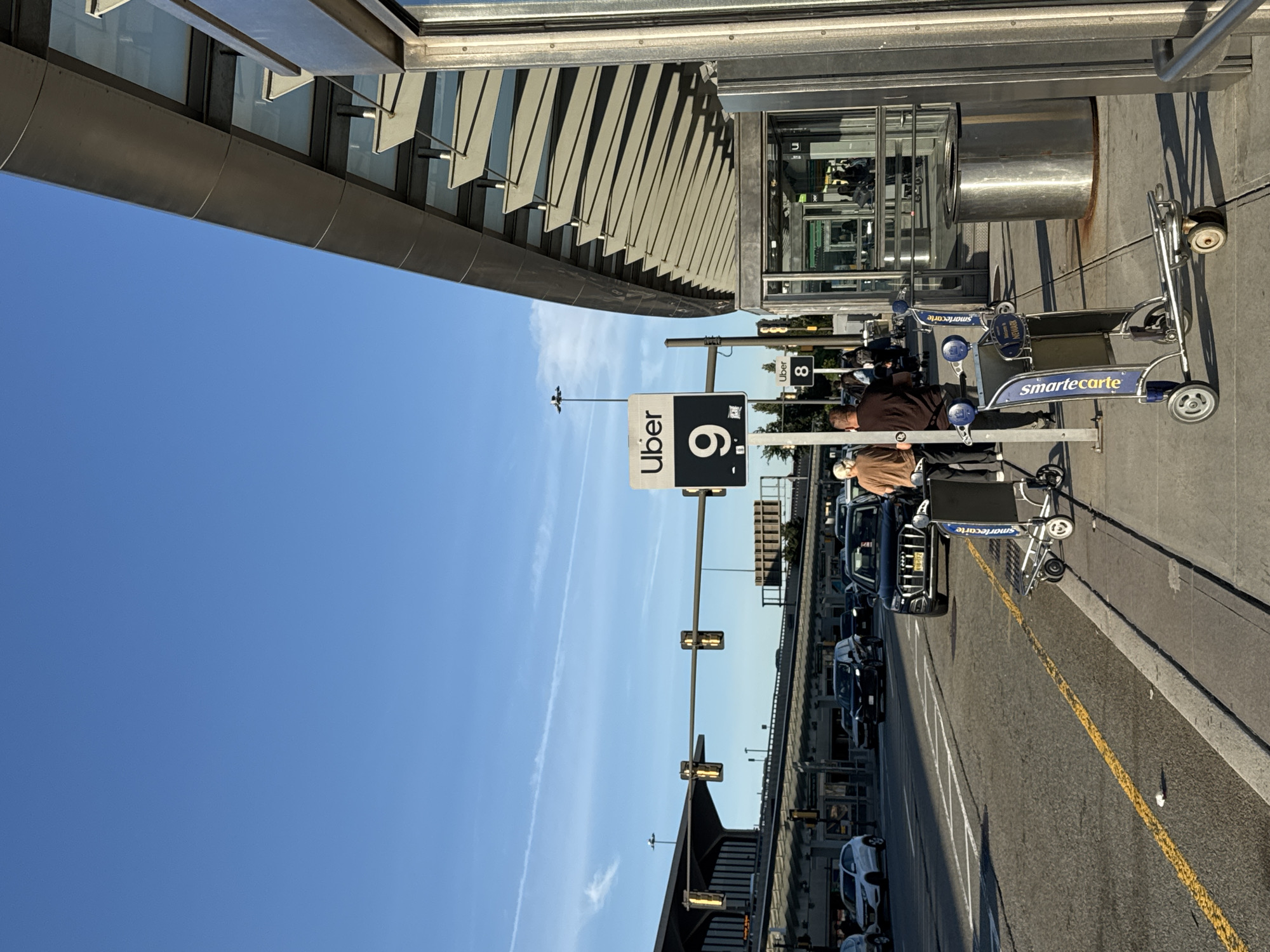 The image depicts a bustling outdoor scene at what appears to be Newark Liberty International Airport, under a bright, clear blue sky with a few wispy clouds, suggesting sunny weather during the late morning or afternoon.
On the left, a modern building facade, likely part of an airport terminal, features metallic horizontal panels and angled sun shades. Adjacent to the building, a paved roadway or drive lane is visible, lined with cars, suggesting a pickup or drop-off area. A black SUV is prominently featured among the vehicles.
Several people are present, mostly appearing to be waiting or moving luggage. One person in a brown jacket stands near a luggage cart, and another in a tan shirt is partially visible behind them.
Key objects include multiple blue and silver SmartCarte luggage carts, some clearly branded with "smartecarte" and "NEWARK." A prominent black sign with white lettering displays "Uber 9," indicating a designated ride-share pickup zone. Further down the road, another white sign with black text reads "Uber 8." Street lights or traffic signals are mounted on poles along the street. The road surface has distinct yellow and white markings.
The scene conveys a typical, functional airport environment, with cues like the "Uber" signs and "NEWARK" on the carts strongly indicating its location in Newark, United States of America.
