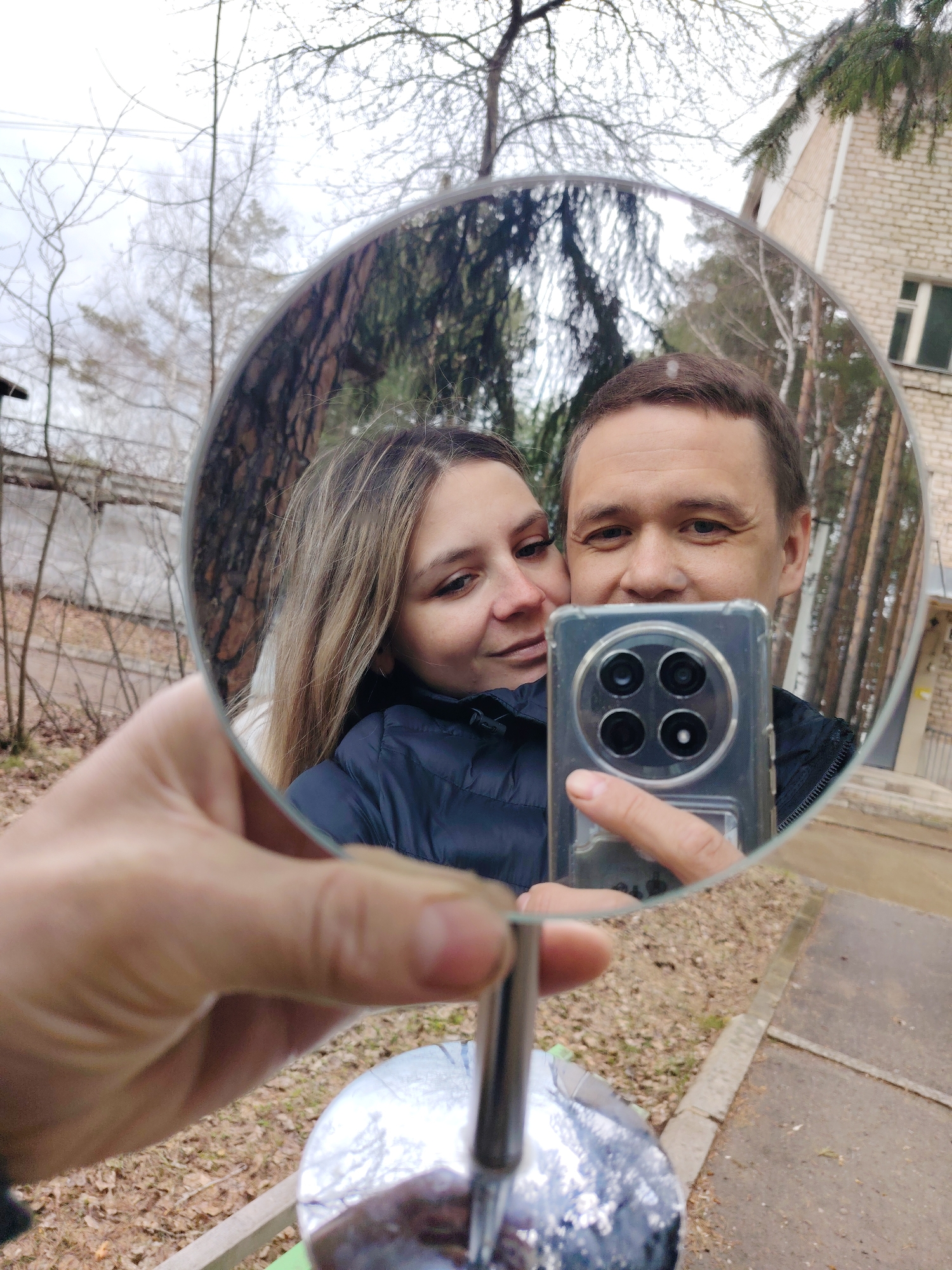 A fair-skinned man and woman are seen in a mirror's reflection, held in someone's left hand, taking a selfie with a phone that has four camera lenses on the back. They are standing in an outdoor environment in Chaikovsky, Russia. The building behind them is made of beige brick with a single window. Various bare and evergreen trees are also present. The mirror's base sits on a light-colored concrete surface with brown leaves and debris scattered around it.