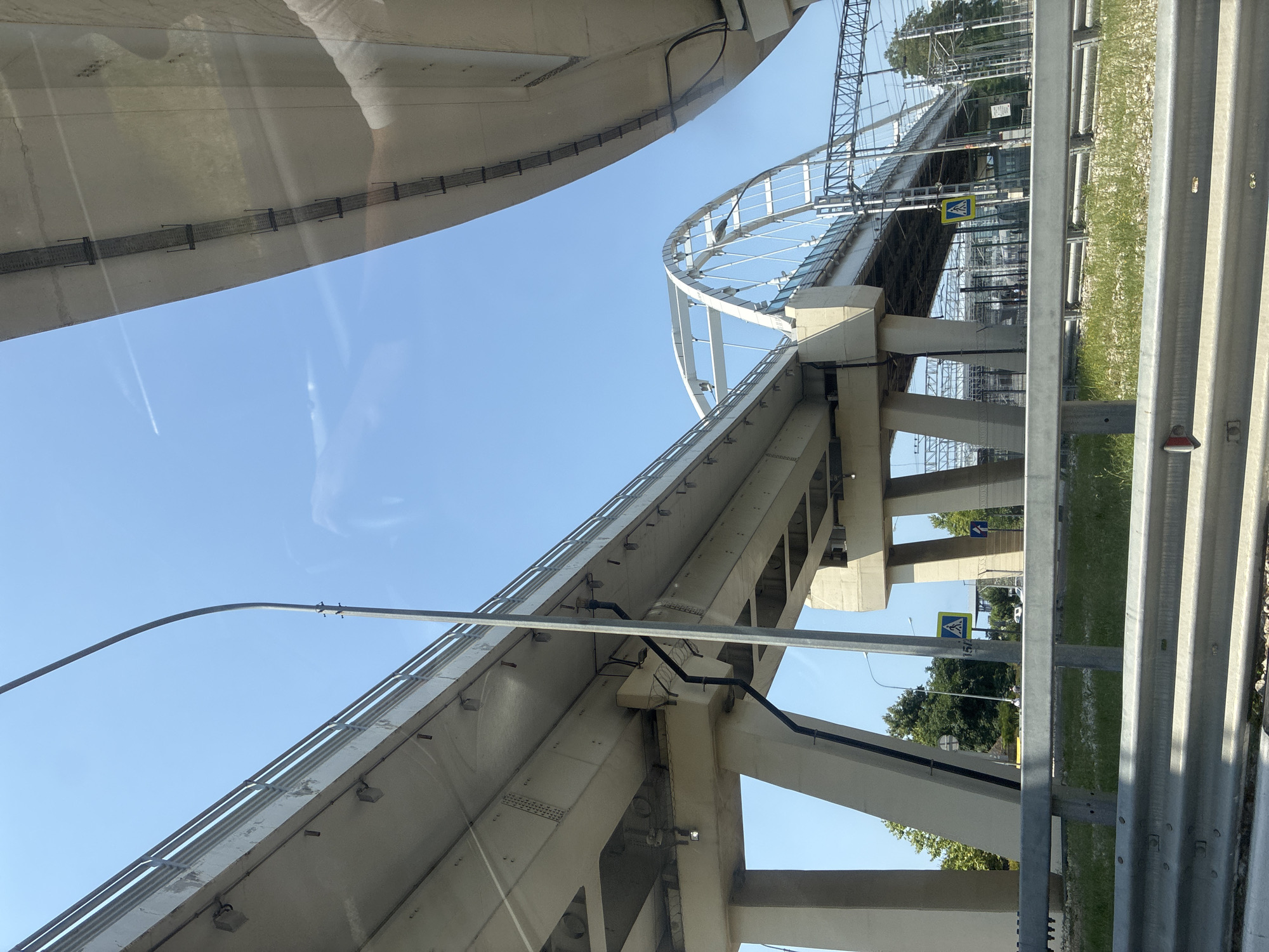 The image captures a view of elevated roadways and a bridge structure against a clear blue sky. In the foreground, a silver metal guardrail separates the viewer from a patch of grass and gravel. Behind the guardrail, large concrete pillars support an elevated roadway that curves towards the right side of the frame. Another elevated structure, appearing to be a bridge with an arched design, is visible further in the distance, with overhead power lines and railway tracks underneath. The scene appears to be an urban or suburban environment, likely a transportation hub. A yellow pedestrian crossing sign is visible on the left side of the image. Given the context of Sochi, Russia, this could be a view of one of the many overpasses or elevated sections of the road infrastructure built for the city, possibly near a railway station or a major intersection. The lighting suggests it is daytime, with the sun casting shadows. No people are clearly identifiable in the frame, though there might be distant figures or vehicles on the roads. The overall impression is one of modern infrastructure within a relatively clear, open environment.