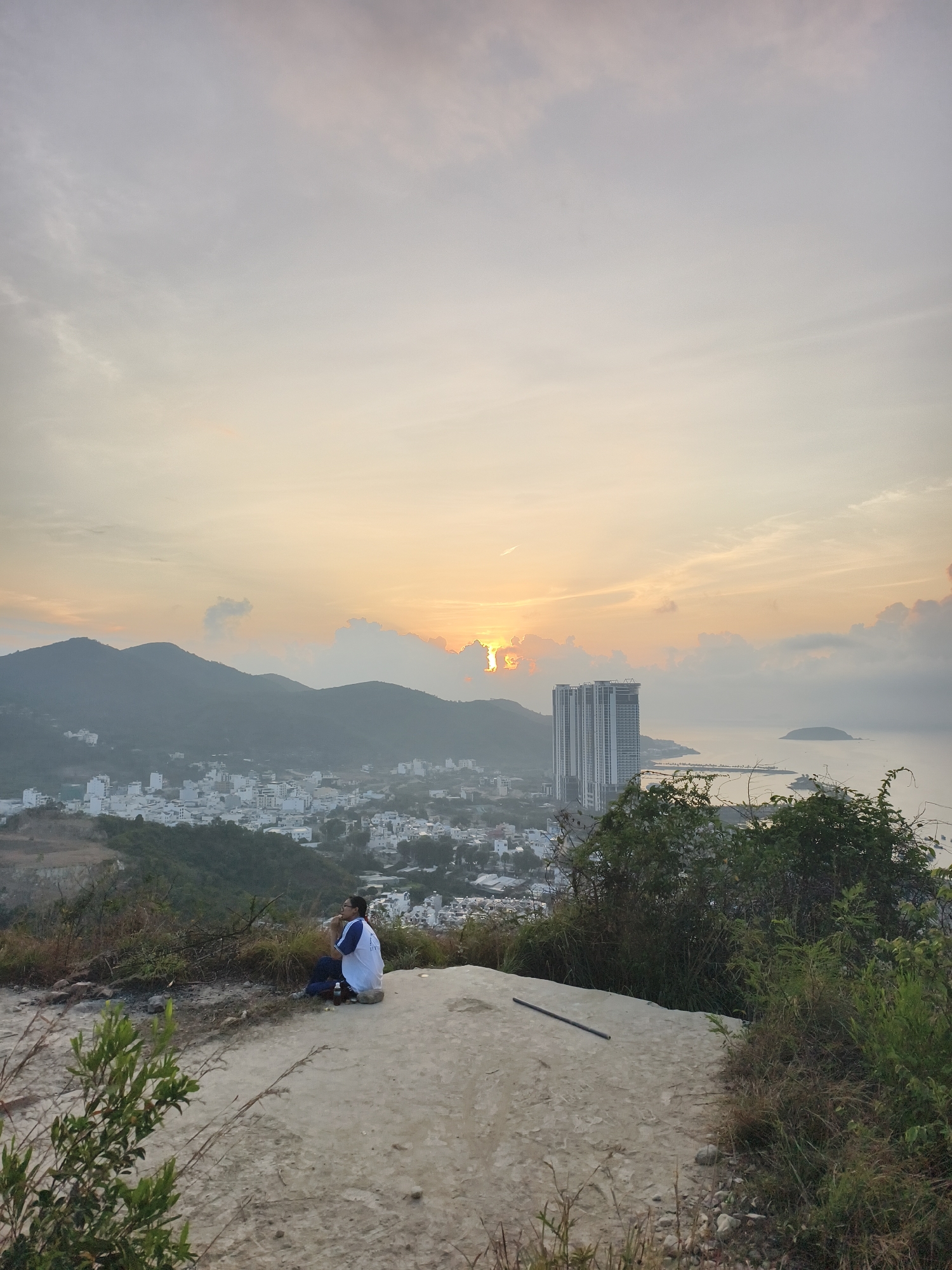 The image presents a serene dawn or sunrise scene captured from an elevated perspective overlooking a coastal area in Bac Nha Trang Ward, Vietnam. In the foreground, a person, appearing to be male and dressed in a white and blue shirt with dark pants, sits on a patch of dry, light-colored ground. He is facing away from the camera, gazing towards the expansive view. Sparse green foliage and dry brush line the edges of the immediate foreground, and a dark, slender object lies on the ground near the seated figure.
The mid-ground reveals a city sprawling between a range of dark mountains and the sea. Numerous white and light-colored buildings characterize the urban landscape. Closer to the water, a cluster of tall, modern high-rise buildings stands out prominently. Beyond the city, the calm sea stretches into the distance, with a small, indistinct island visible through a slight haze.
The sky is bathed in the soft, warm hues of sunrise, with the sun's golden light peeking through and illuminating soft clouds along the horizon. The upper sky transitions into lighter grays and blues, creating a gentle, diffused light over the entire scene. The overall atmosphere is one of peaceful contemplation, as the individual witnesses the start of the day over the scenic Vietnamese coast. No visible text is present in the image.