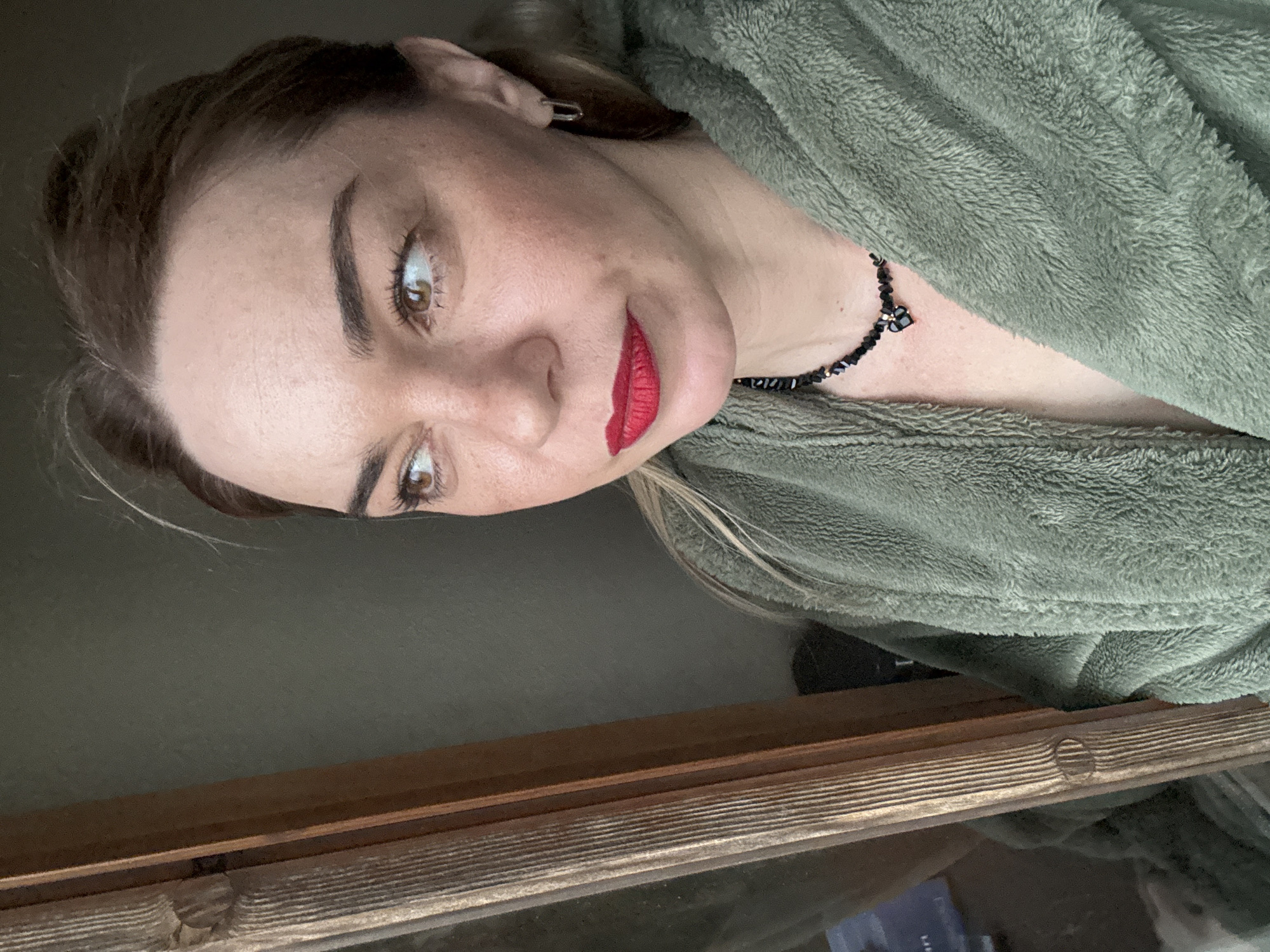 A woman is shown in a selfie. She is wearing a green robe and a black beaded necklace. Her lips are red. She appears to be indoors. The background includes a wooden structure and a blurry dark surface. The location is not possible to determine from the image.