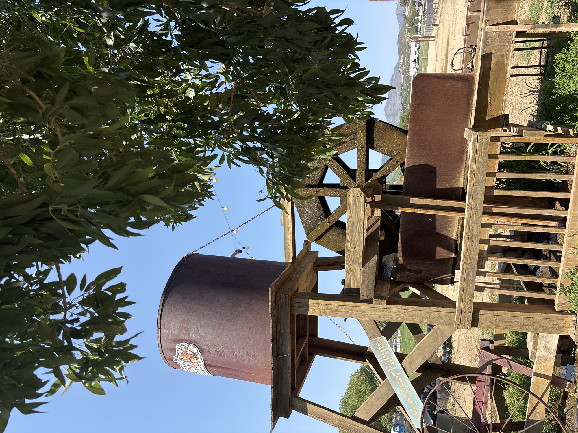 The image depicts a rustic wooden water wheel and associated structure, likely an attraction at a farm or park, located outdoors under a clear blue sky with abundant foliage from a tree overhead. The structure is constructed from aged wood and features a large, rusty metal water tank at the top, feeding into the water wheel. The water wheel itself is made of wood with metal buckets. A wooden sign on the structure reads "Mountain Farms." In the background, there are more trees, some fencing, and in the distance, a hilly landscape with buildings and parked vehicles, suggesting a semi-rural or suburban environment. The area around the base of the structure appears to be sandy or dirt ground with some greenery. The scene is brightly lit by sunlight, casting shadows. There are no people visible in the image.