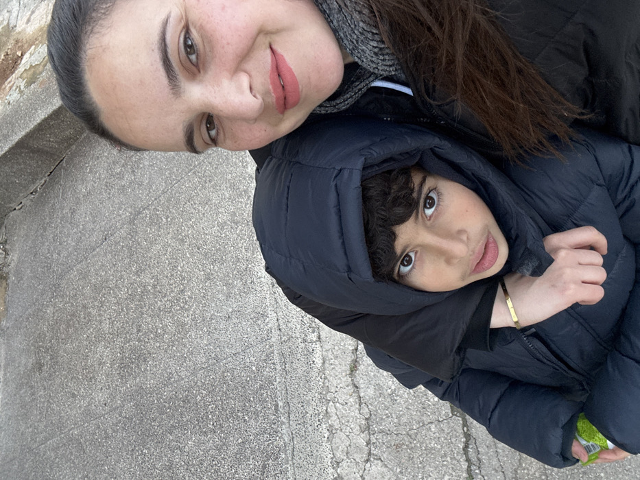 An overhead, diagonal shot captures an adult woman and a child posing closely together outdoors on a grey, textured ground surface. The image is oriented so that the subjects' heads are angled upwards from the bottom left towards the top right.
The adult woman, positioned in the upper half of the frame, has fair skin with some freckles, light brown eyes, and dark eyebrows. Her dark hair appears to be pulled back. She is wearing a dark outer jacket, a grey patterned scarf around her neck, and a light pink lipstick. She is smiling subtly with her eyes and looking towards the camera.
The child, who appears to be a boy, is situated below and to the right of the woman, leaning against her. He has dark, curly hair visible under a dark blue or black puffy winter jacket with the hood up, framing his face. He has large, dark brown eyes and is looking directly at the camera with a neutral expression. On his left wrist, a thin, gold-colored bracelet is visible. A small, crumpled green and white object, possibly a wrapper, is partially seen in the child's hand within the jacket at the very bottom right corner of the frame.
The background consists primarily of a rough, grey concrete or asphalt surface, suggesting an outdoor location like a sidewalk or paved area. The lighting indicates daylight, with soft illumination and no harsh shadows, potentially suggesting an overcast day. The heavy winter jackets worn by both individuals indicate cool or cold weather. No discernible text is visible in the image.