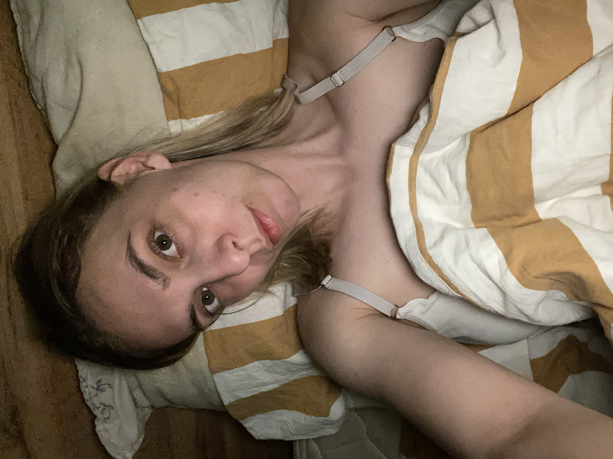 A woman is lying in a bed covered with a striped yellow and white blanket. She is wearing a light-colored top with straps. Her head rests on a pillow. The image appears to be taken indoors. There is no indication of the location being Chaikovsky, Russia, in the image.