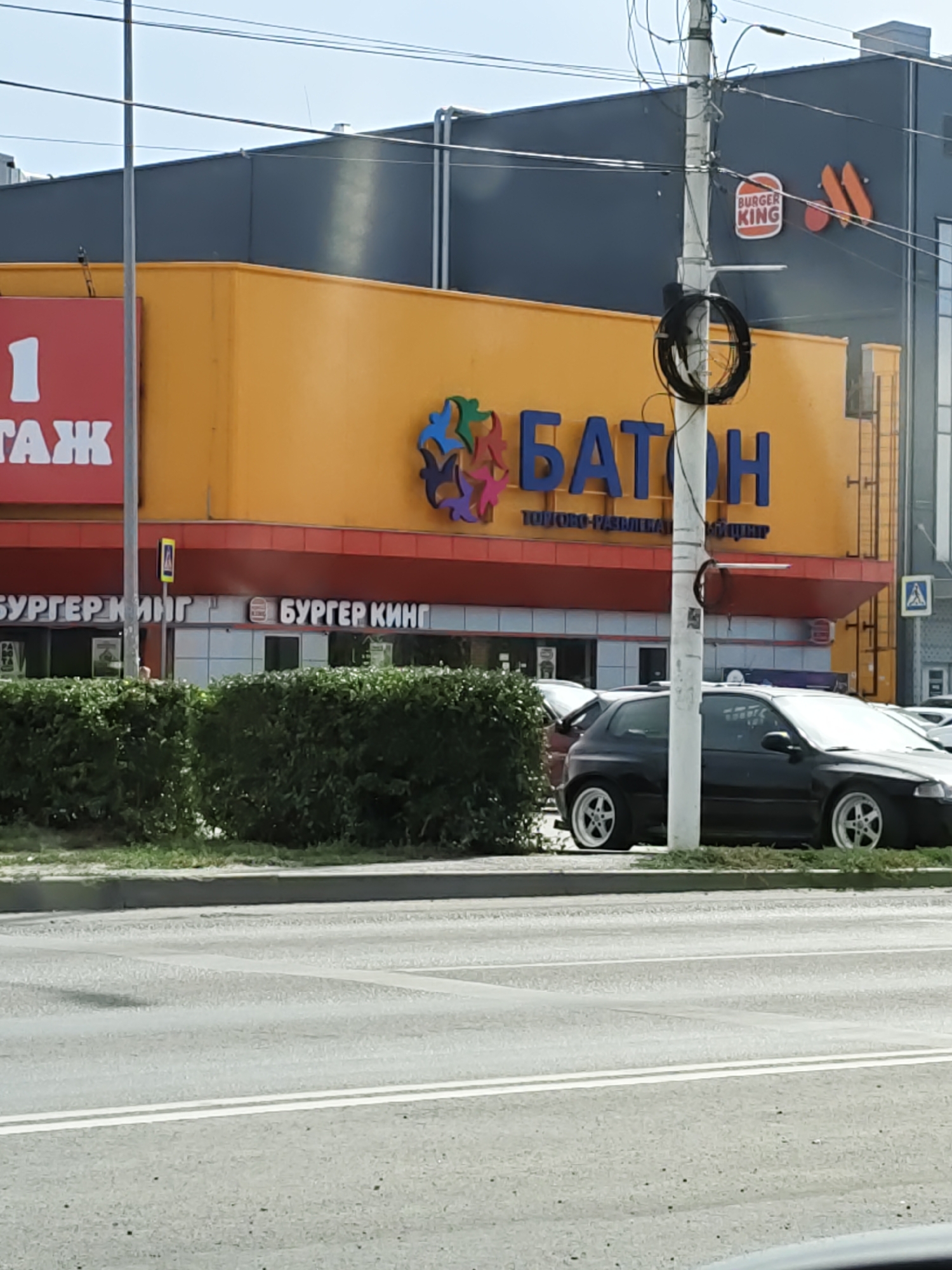 The image shows the exterior of a commercial building with a bright yellow facade. A large blue sign with the word "БАТОН" (Baton) is prominently displayed. Below this, smaller text reads "ТОРГОВО-РАЗВЛЕКАТЕЛЬНЫЙ ЦЕНТР" (Shopping and Entertainment Center). To the left of the "БАТОН" sign, there is a red sign with a white "1" and the letters "ТАЖ" (Floor). To the right, a Burger King logo is visible on a dark blue building facade, and another Burger King sign in Russian ("БУРГЕР КИНГ") is seen on the lower level of the yellow building. A black hatchback car is parked in front of the building, with the lower portion of a second car visible behind it. Green bushes line the sidewalk. A traffic light pole with visible wires is positioned in the foreground. The scene is set on a street with a paved road and white lane markings, indicative of an urban environment. This commercial building complex is located in Novocherkassk, Russia.