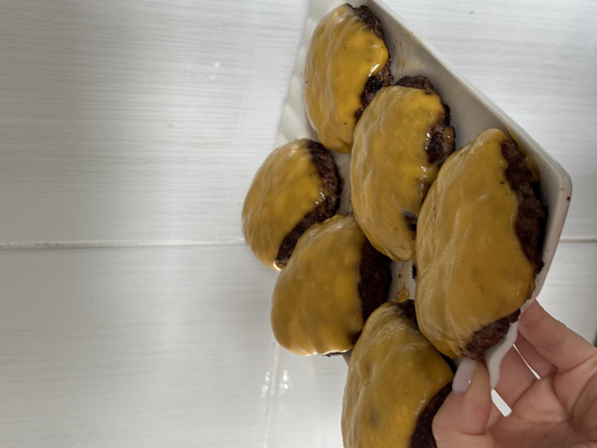Six cheeseburger patties covered in melted cheese are arranged on a rectangular white plate. The patties are circular and have a dark brown color, with the melted cheese a vibrant yellow-orange. The plate is positioned on a white surface that appears to be a countertop or shelf, with a textured white wall in the background. The lighting is bright, casting soft shadows. A human hand with a light-colored fingernail is visible on the left side of the image, gently holding the edge of the plate. The scene appears to be in a domestic kitchen setting. There is no information in the image that would allow for determination of its location within the city of Поназырево, Russia.