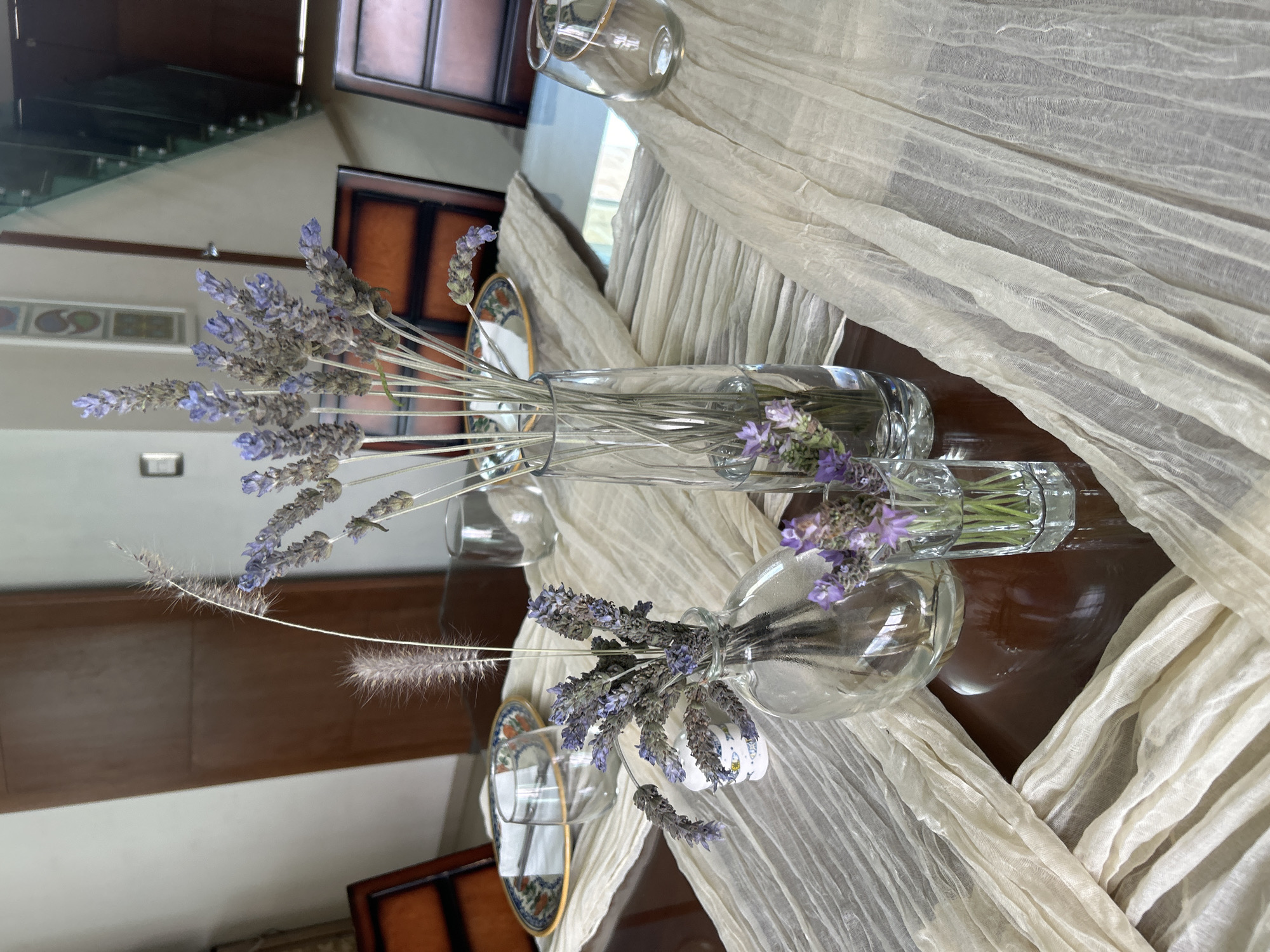 The image shows a table setting indoors. Three transparent glass vases are placed on a beige, draped cloth. Each vase holds sprigs of lavender. One vase also contains feathery grasses. To the left of the vases, there are small plates with a blue and gold rim. The table surface is dark brown wood. In the background, reflective surfaces suggest the interior of a home with stairs and architectural details. The location in Barrio de Nuevo León, Mexico is not verifiable from the content of the image.