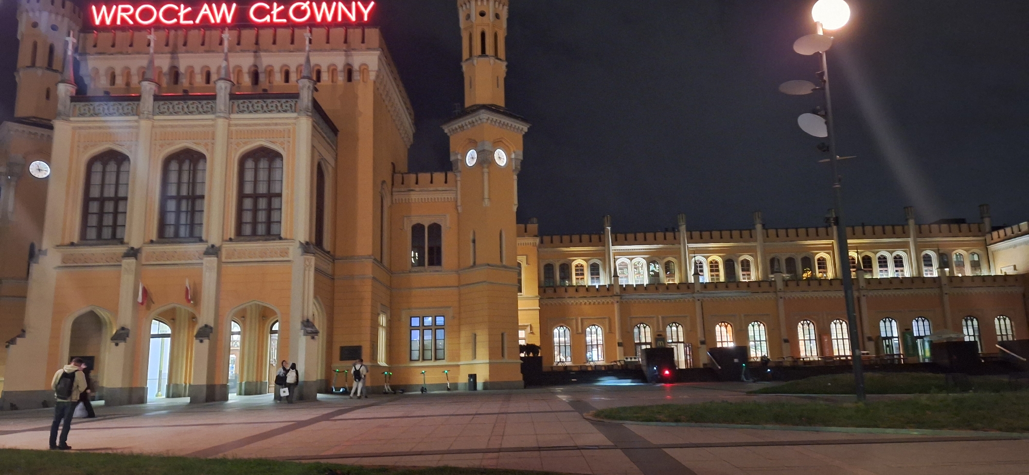 The image captures the Wrocław Główny railway station in Wrocław, Poland, at night. The station's main building, illuminated by warm light, is a grand ochre-colored structure with Gothic architectural elements, including pointed arch windows and crenellated parapets. A prominent clock tower, also ochre, stands to the right of the main building, displaying two clocks. Red neon lights spell out "WROCŁAW GŁÓWNY" across the top of the building.
In the foreground, a paved plaza with patches of grass is visible. A tall lamppost with a glowing orb at the top stands on the right side of the frame, casting a beam of light upwards. Several people are present on the plaza. On the left, a man wearing a light-colored hoodie and a backpack stands facing away from the camera, looking towards the station entrance. Two individuals are standing near an entrance on the left, one with a bag. Further to the right, a person in a light-colored outfit stands near several parked electric scooters. More people are visible in the distance, near the illuminated windows of the station's longer wing. The sky is dark, indicating it is nighttime.