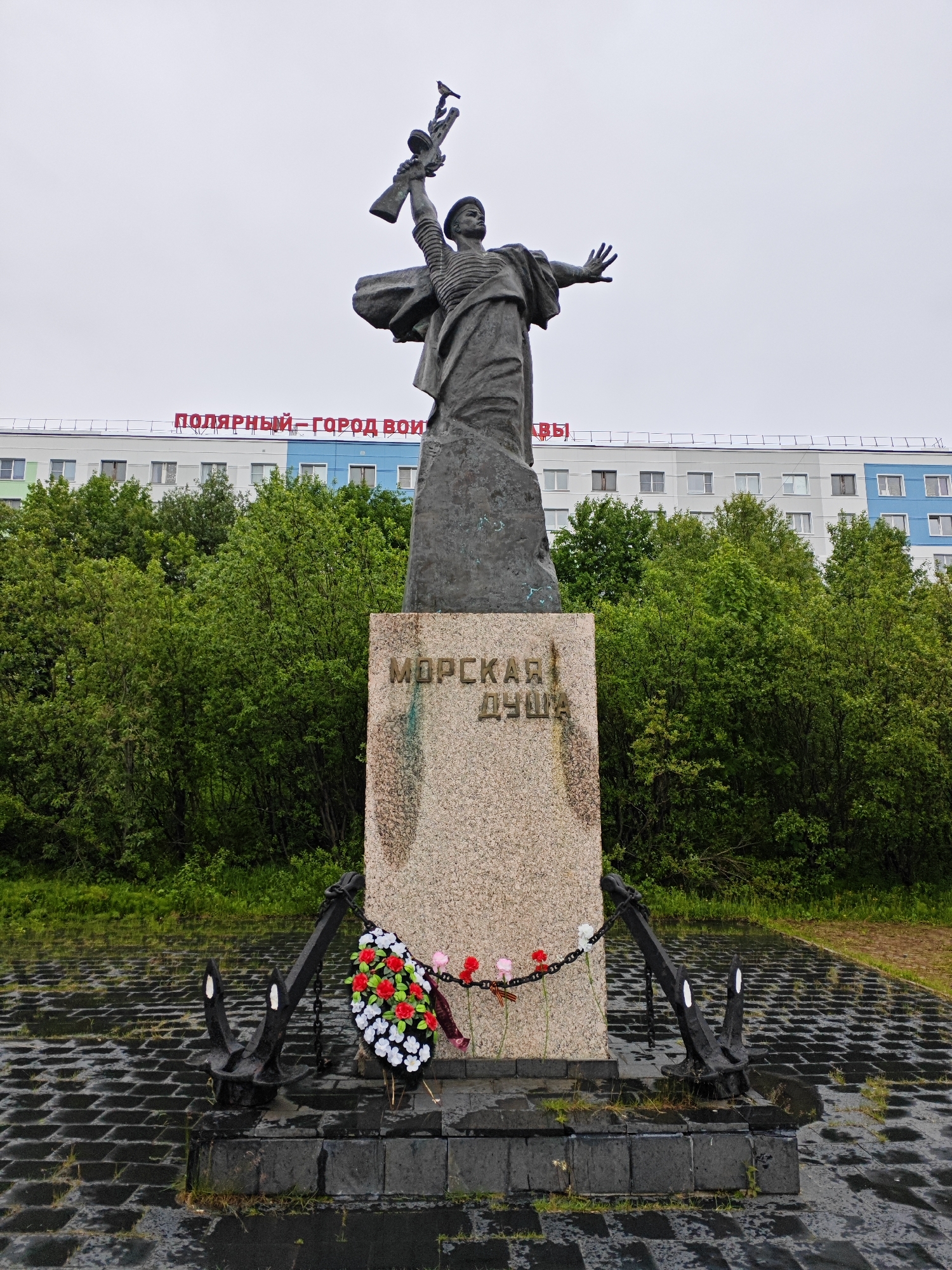 This media file features a statue of a sailor holding a rifle, with his arm outstretched, standing on a granite pedestal. The pedestal is adorned with a wreath of red and white flowers and is flanked by two large black anchors. The statue and monument are situated on a wet, dark paved area, suggesting it has recently rained.
Behind the monument, a building with a light-colored facade is visible, featuring windows and balconies. Above the building, red Cyrillic lettering spells out "ПОЛЯРНЫЙ – ГОРОД ВОИНЫ", which translates to "Polar – City of Warriors". Lush green trees form a dense backdrop behind the building and to the sides of the monument. The sky is overcast and a dull gray, indicating cloudy or rainy weather. The overall scene depicts a solemn memorial, likely in the city of Polyarny, Russia, a place recognized as a "City of Warriors." The wet ground and subdued lighting contribute to a somber and reflective atmosphere.