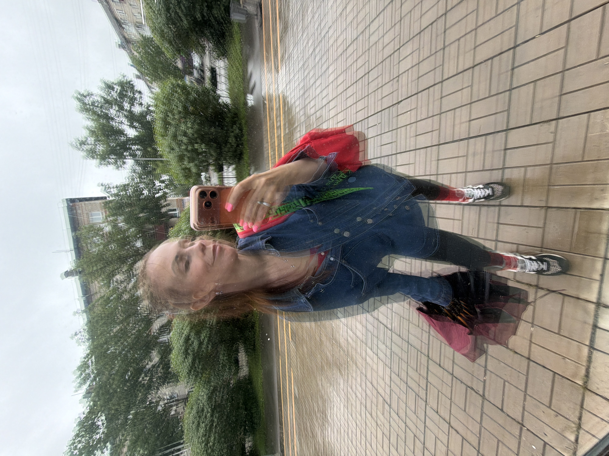 A young woman with auburn hair is reflected in a wet paved surface, suggesting a city street in Birobidzhan, Russia, likely during or after rainfall given the reflections and the closed umbrella visible on the ground. The woman is wearing a denim jacket over a red top, black leggings with red-striped socks, and athletic shoes. She is holding a smartphone in her right hand, angled to take a selfie, and has a red bag with a green strap slung over her shoulder. The background of the reflection shows buildings and trees, consistent with an urban environment. The wet pavement creates distorted reflections of the woman and her surroundings, indicating a damp, overcast day.