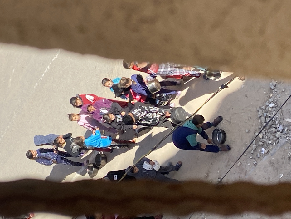An overhead view in Khan Yunis, Palestinian Territory, shows approximately twenty individuals, primarily children and adolescents, gathered on a hard-packed dirt or paved surface. A blurred, brown vertical structure partially obstructs the scene from the right and left, with two dark cables traversing the lower foreground. Strong sunlight illuminates the area, casting prominent shadows. The individuals are dressed in varied casual clothing, including hoodies and long pants, in colors such as blue, purple, pink, red, and black. Many are holding or standing near metal pots, buckets, and bowls of various sizes. In the lower center, one child in a blue and teal tracksuit is bent over a black bucket on the ground. The arrangement of the individuals and their containers suggests a queue or assembly for the collection of a resource.