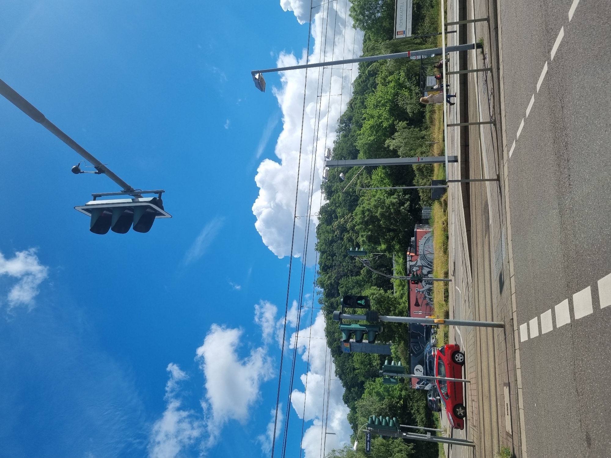 The image shows a street scene in Saarbrücken, Germany, on a bright, sunny day with a blue sky and scattered white clouds. In the foreground, there is an asphalt road with white dashed lines indicating a crosswalk. A red car is visible driving on the road. Across the street, there are tram tracks and a pedestrian area with a few people walking. A traffic light with a green bicycle signal is prominent. Behind the street, a densely wooded hill rises, with some buildings and structures partially visible amongst the trees. Overhead, power lines stretch across the scene, supported by poles. A traffic light hangs suspended from a long arm over the intersection. A street sign that reads "S RÖMERKASTELL" is visible on the right side of the image. The overall impression is of a typical urban environment with a focus on transportation infrastructure and green surroundings.
