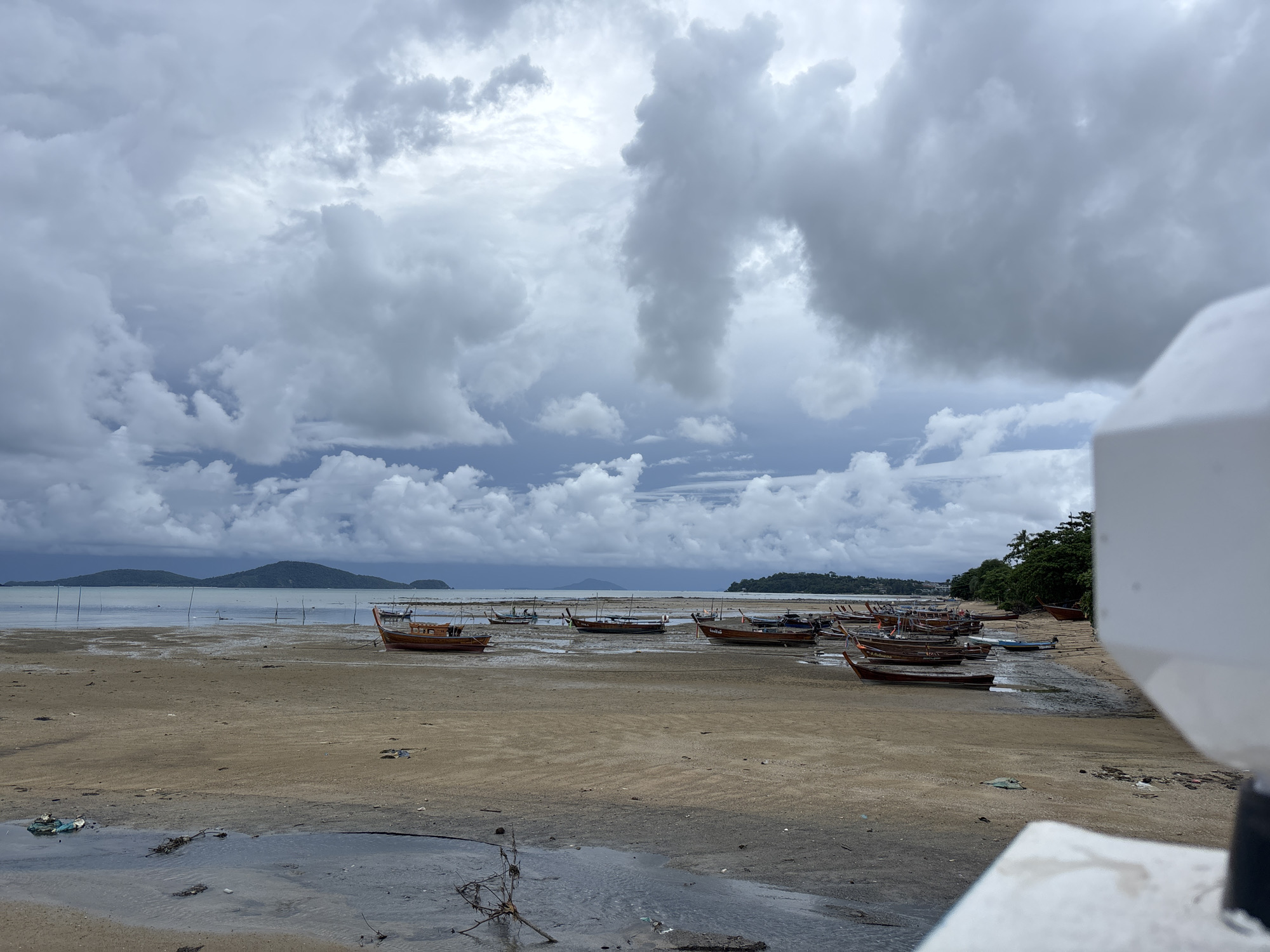 The image captures a beach scene in Rawai, Thailand, under a dramatic, overcast sky filled with dark, heavy clouds. Numerous longtail boats are beached on the sandy shore, some partially submerged in shallow water or mudflats. The tide appears to be out, exposing the wet sand and leaving pools of water. In the distance, lush green islands rise from the sea, silhouetted against the turbulent sky. The foreground shows a muddy, wet area with debris scattered on the sand, including plastic waste and branches. A white, out-of-focus object, possibly a railing or structure, is visible in the lower right corner, partially obscuring the view. The overall atmosphere is somber and somewhat desolate, suggesting an impending storm or the aftermath of one.