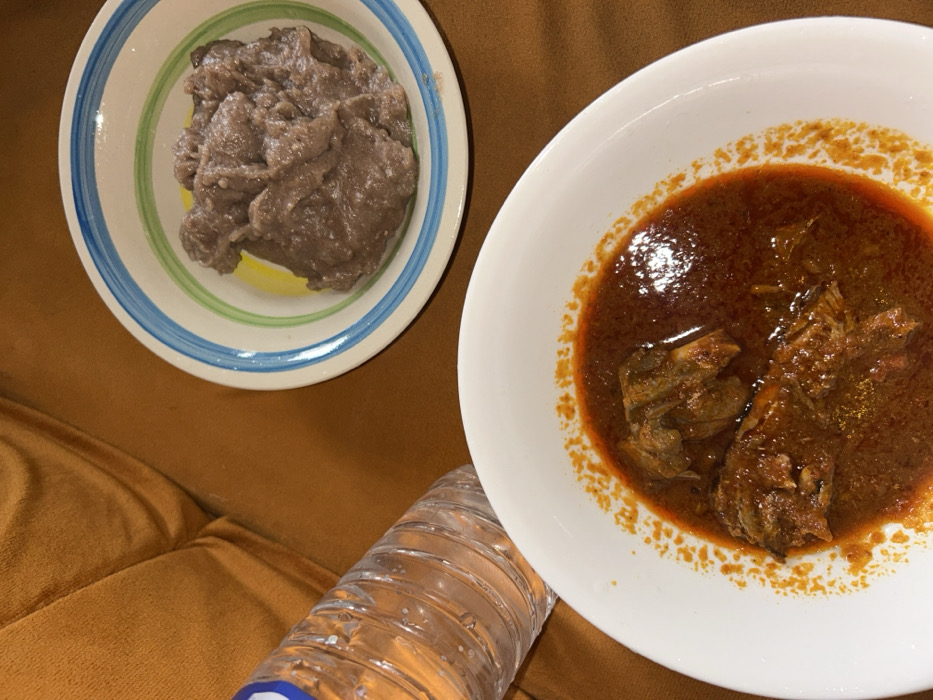 In Egbe, Nigeria, a meal is displayed on a brown, upholstered surface. In the foreground, a white bowl contains a red-orange stew with several pieces of what appears to be meat or fish. A clear plastic water bottle, partially filled with water and featuring a blue cap, rests to the left of the stew bowl. In the background, a second white bowl, adorned with blue and green stripes, holds a quantity of brownish-grey, dense solid food. No individuals are visible in the frame.
