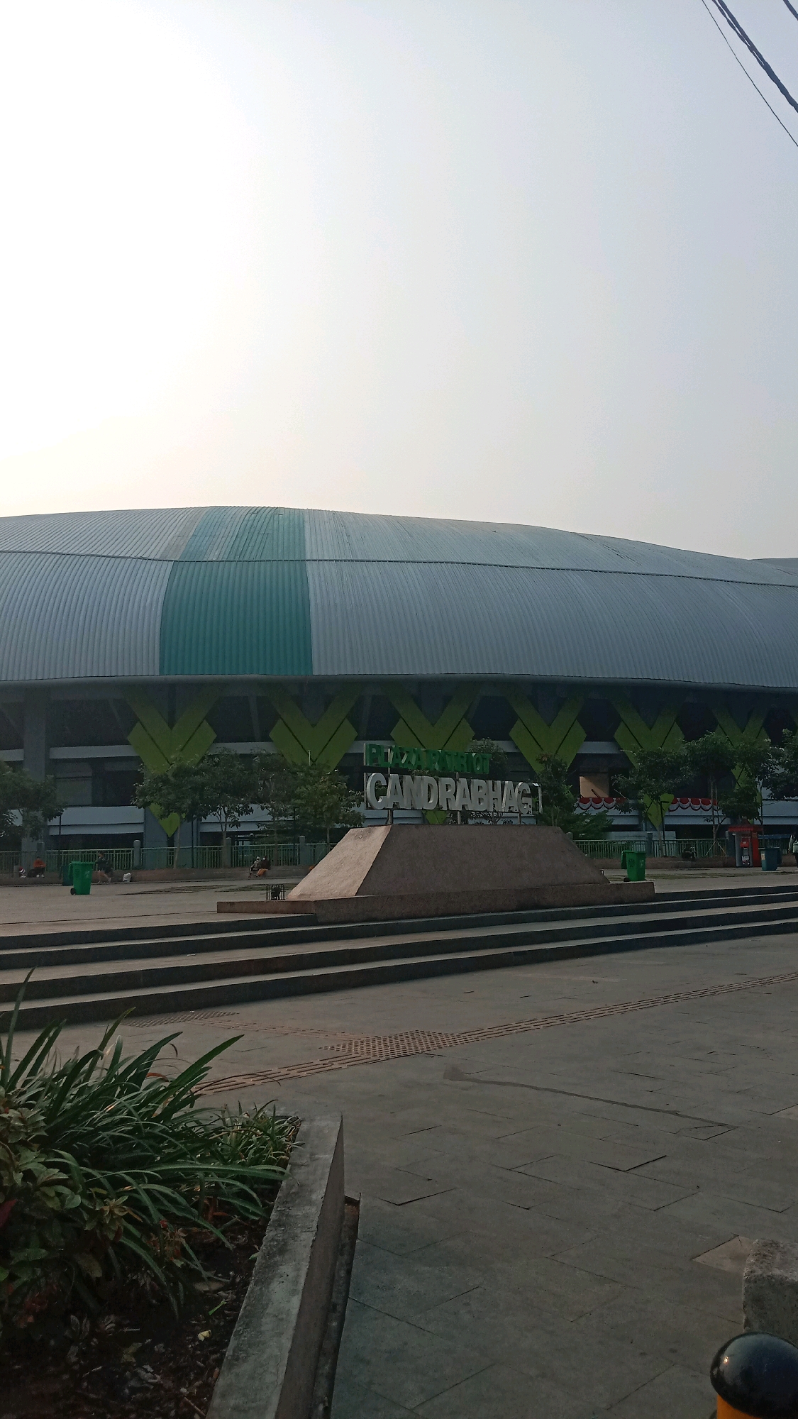 A wide-angle shot captures the facade of the Patriot Chandrabhaga stadium in Bekasi, Indonesia, under a hazy sky. The stadium's prominent feature is its arched, silver-colored roof with a wide, horizontal strip of green and white banding. Below the roofline, a dark band is visible, broken by repeating angular patterns in bright green. In the foreground, a low, stepped concrete platform leads up to a monument with the words "PATRIOT CANDRABHAGA" inscribed in large, raised silver letters. Several small trees with green foliage are planted in front of the stadium's lower levels, and some trash bins are visible. The ground in front of the monument is paved with gray squares, and a textured tactile paving strip is present. In the lower left of the frame, a planter with lush green plants is visible. A few individuals are scattered in the background, sitting or walking, and a green trash can is near one of them. The overall scene suggests an outdoor public space in front of a large sports venue.