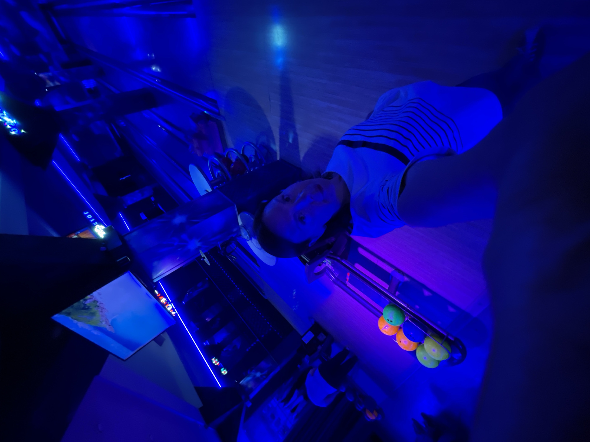 A low-angle shot shows a person wearing a striped white shirt, standing inside a bowling alley with blue lighting. The person is in front of a ball return area, with multiple bowling balls visible. Beyond, the lane is partially visible with a scoring screen displaying an image. "SOCHI" is visible on the screen.