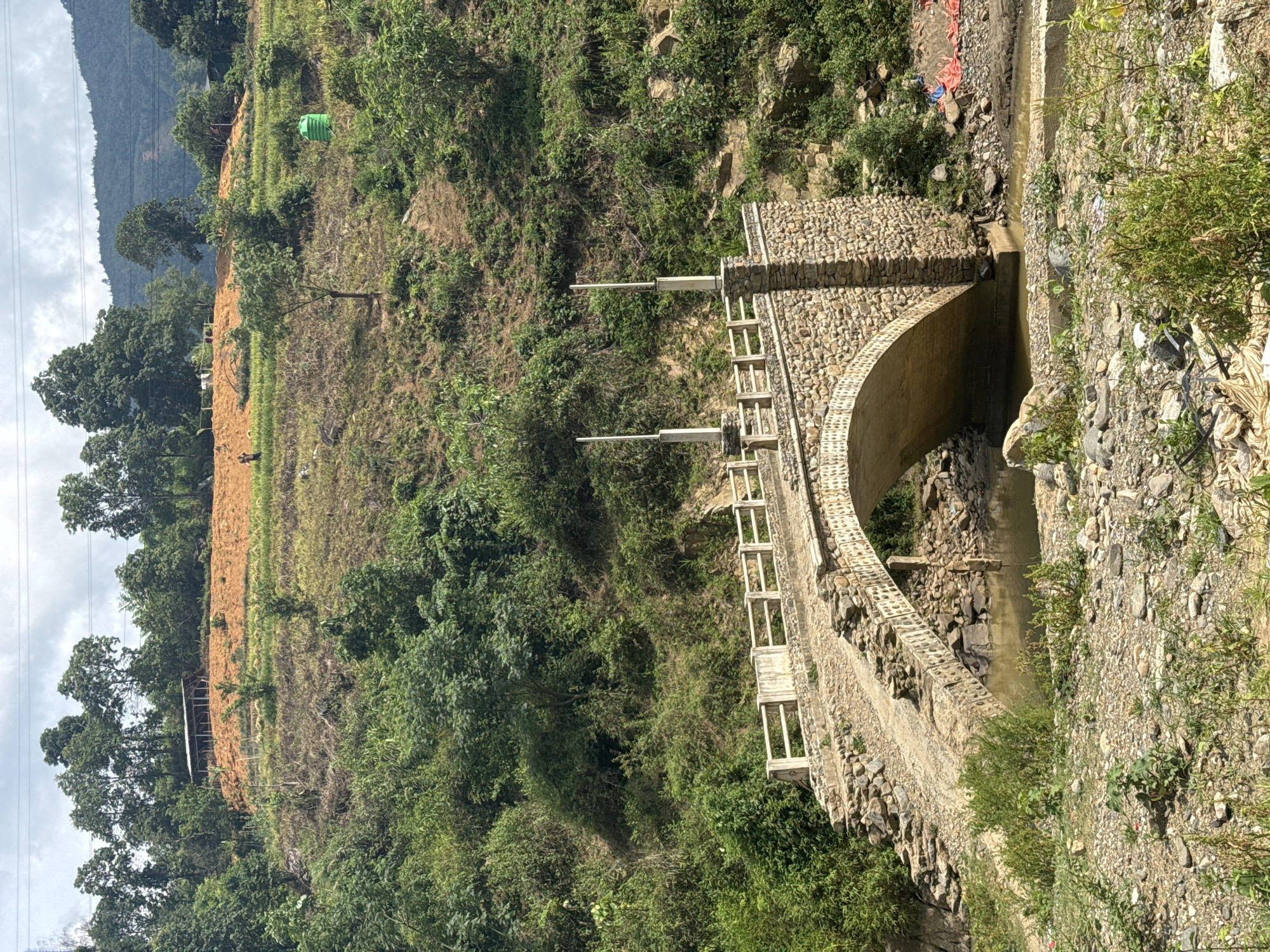 This media file presents a vibrant, mountainous landscape in Khao Mang, Vietnam, featuring a stone arch bridge or aqueduct as a prominent subject. The scene is captured during daytime under an overcast sky, providing bright, diffused light.
The central feature is a sturdy, well-constructed stone arch bridge with low concrete railings and two taller, slender concrete posts extending upwards from its top. This structure spans across a narrow, shallow stream with rocky banks, flowing from the right towards the left side of the frame. The stream appears to be a natural watercourse, with some scattered debris and plastic bags visible along its banks on the right side.
To the left of the bridge, a steep hillside is visible, partially covered with lush green trees and dense foliage, and partially cleared with exposed brown earth, possibly indicating agricultural activity or recent land disturbance. A bright green plastic water tank is situated on this cleared hillside. In the mid-ground of this left section, small, indistinct structures or shelters can be discerned amidst trees. Further in the background, a large mountain range is silhouetted against the cloudy sky. Power lines are faintly visible against the sky on the far left.
To the right of the bridge, the terrain is also hilly and densely covered with various types of green vegetation, including trees, bushes, and vines, suggesting a wilder, less disturbed environment compared to the left side. No identifiable people or visible text are present in the images.