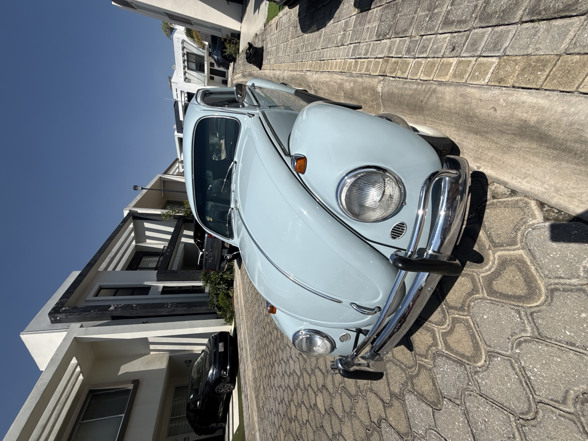 A light blue Volkswagen Beetle is parked on a paved street in Santa Clara Ocoyucan, Mexico. The car is angled towards the viewer and parked next to a modern-style building with white walls and dark accents. A black sedan is also parked nearby, partially obscured by the building. The street is made of patterned paving stones, and the sky is clear and blue.