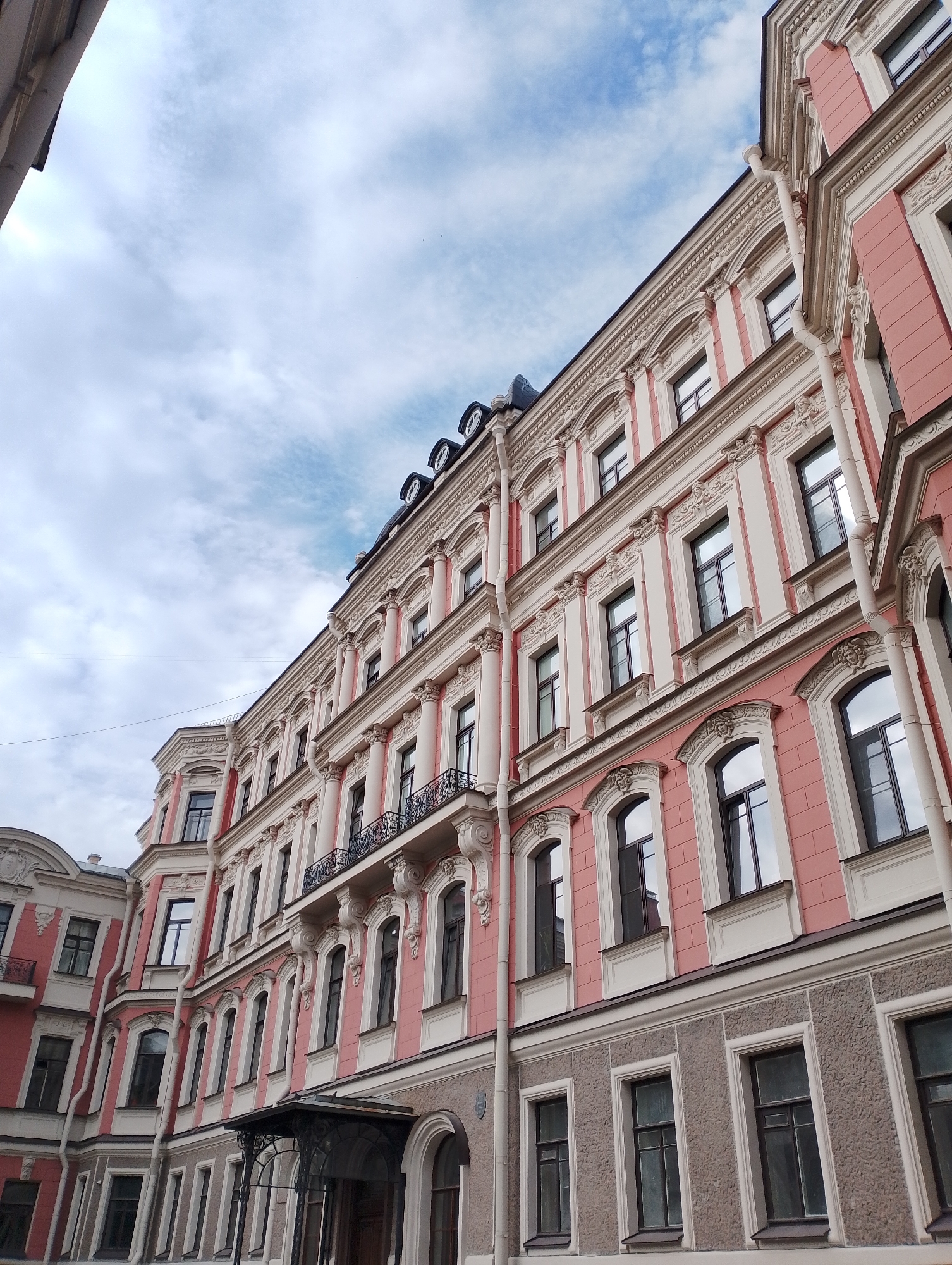 A low-angle, eye-level shot captures the ornate facade of a multi-story building in Saint Petersburg, Russia, under a cloudy sky. The building's exterior is a striking combination of pale pink and cream-colored architectural elements, featuring decorative molding, columns, arched windows, and balconies with intricate wrought-iron railings. The lower level of the building is constructed with a rough, grey stone. The sky above is a pale blue with scattered, wispy white clouds. The perspective emphasizes the height and grandeur of the architecture, with the building extending upwards and to the right of the frame.