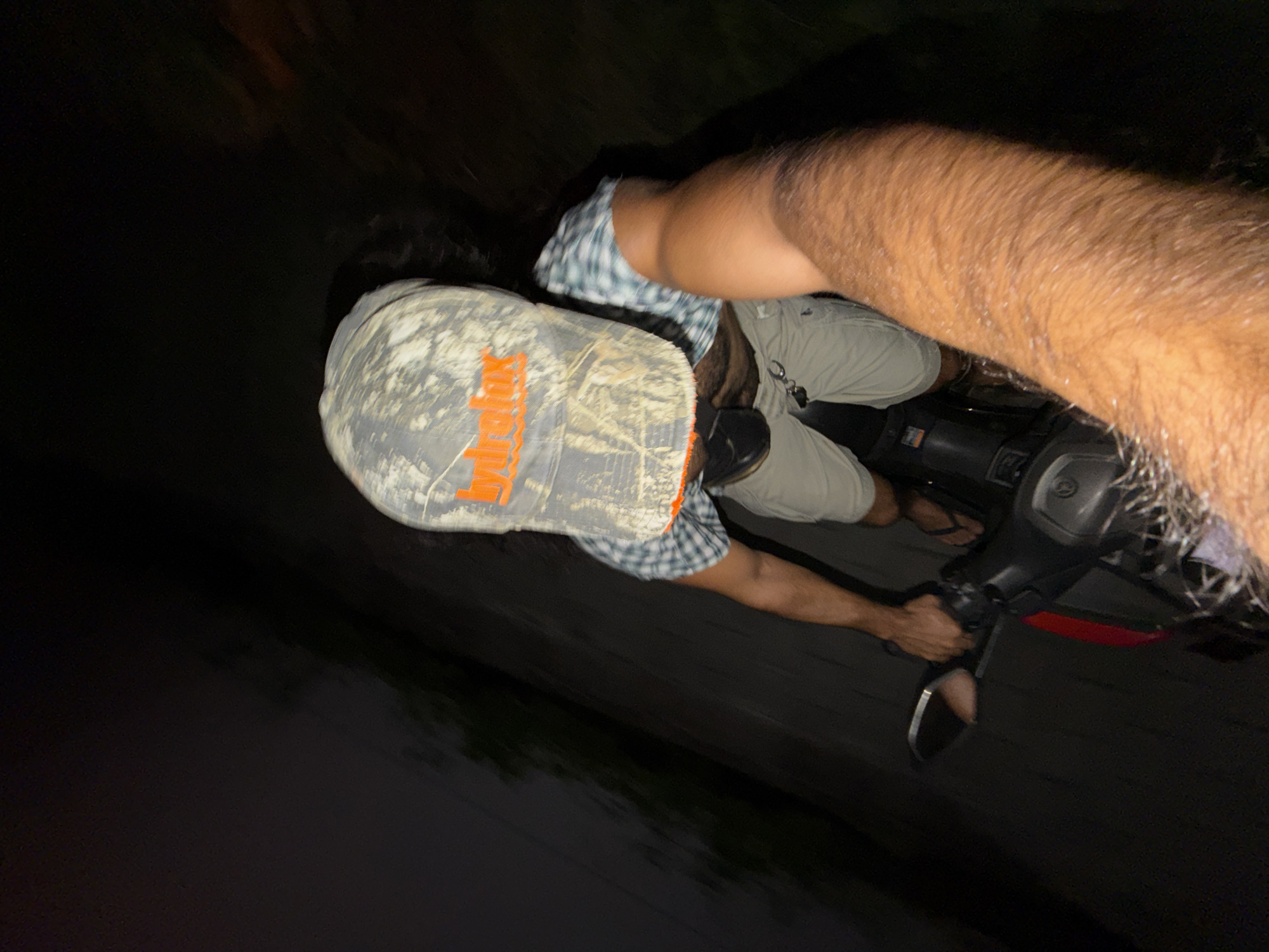 A male is operating a moped at night. The man is wearing a camouflage hat, plaid shirt, khaki shorts, and flip-flops. The moped has a red reflector. The photograph appears to be taken by the operator of the moped. The location is North Kuta, Indonesia.