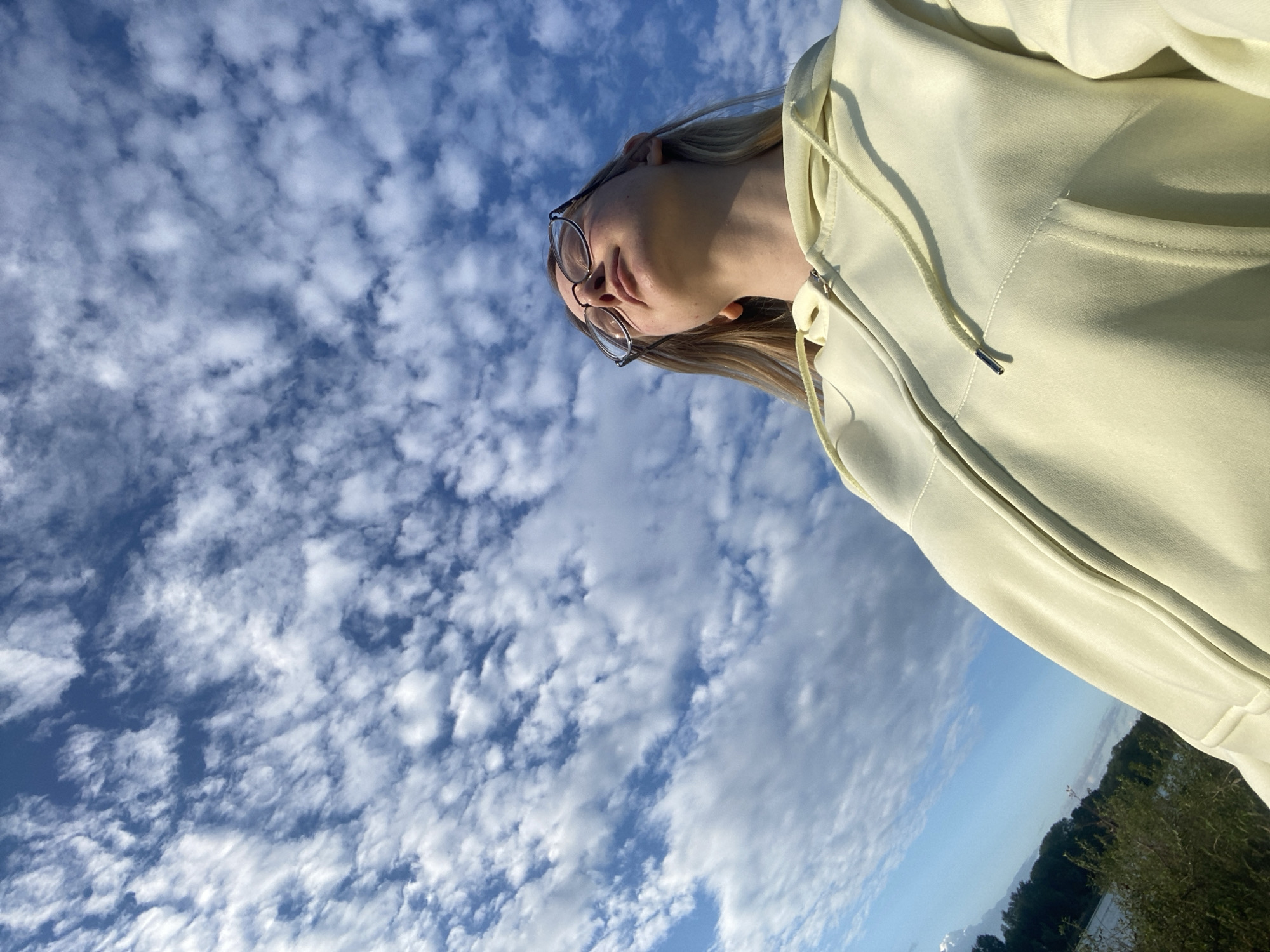 A young woman wearing glasses and a light yellow hooded sweatshirt is captured in a low-angle shot, looking upwards towards a bright blue sky filled with scattered white clouds. Her eyes are closed, and her face is tilted back, suggesting a moment of contemplation or enjoyment of the outdoor environment. The background includes a body of water and a tree-lined shore, indicative of a natural setting. The image was taken in Chaikovsky, Russia, likely near the Kama River, given the landscape depicted. The lighting suggests it is daytime, possibly late afternoon given the angle of the sun.