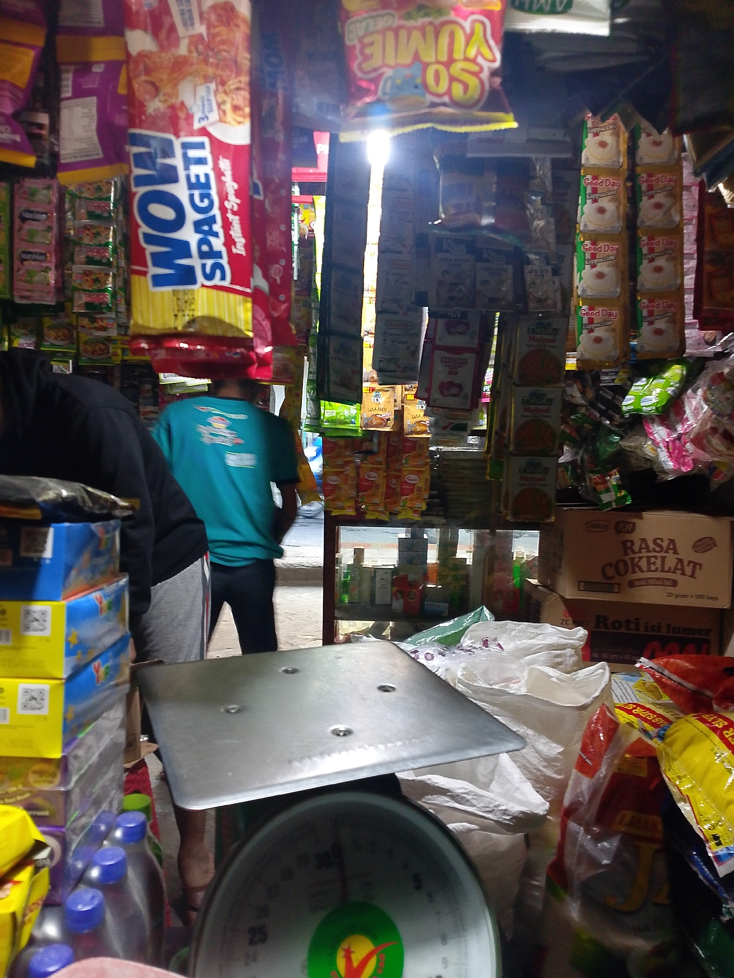 This image captures a view inside a small shop, likely a warung or mini-mart, located in Palembang, Indonesia. The shop is filled with shelves and hanging displays of various packaged food items, including instant noodles, snacks, and beverages. In the foreground, a metal weighing scale is prominently displayed. Behind the scale, two individuals are visible. One person, wearing a dark hoodie and grey sweatpants, is partially obscured by the scale and a stack of blue boxes, suggesting they may be a customer or shop employee. Another individual, wearing a teal t-shirt and dark pants, stands further back in the shop, facing away from the camera. The lighting suggests it might be daytime, with sunlight filtering in from an opening, creating some bright spots and shadows. The overall impression is of a typical, bustling neighborhood store in Indonesia.