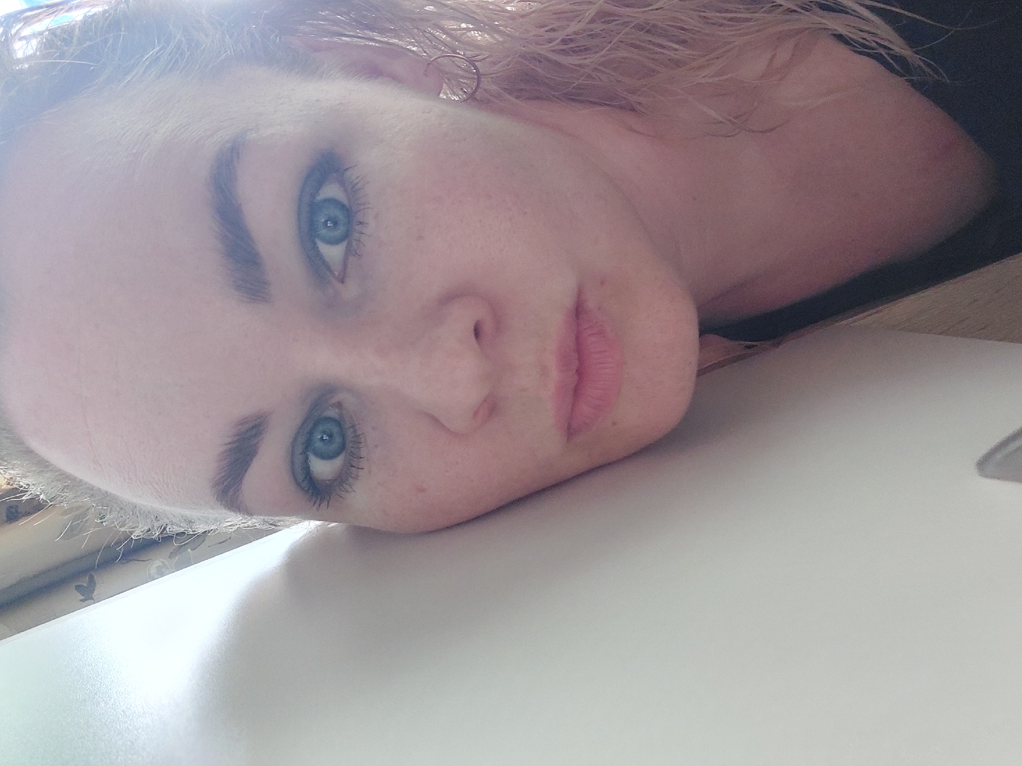 This is a close-up, tilted selfie of a young Caucasian woman. She has fair skin with freckles, blue eyes, dark eyebrows, and light blonde hair. She is wearing a small, hoop earring in her left ear and what appears to be a dark top. Her right cheek rests on a white surface, possibly a desk or table, which dominates the lower half of the frame. The background is blurred, but appears to include a wooden surface and some patterned fabric. The lighting suggests it could be daytime, with a soft light illuminating her face. The woman's expression is neutral, with her lips slightly parted. There is no visible text in the image.