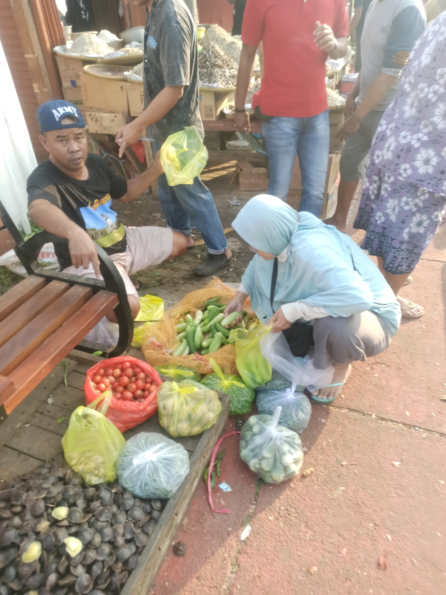The image depicts an outdoor market scene in Cikarang, Indonesia. In the foreground, a man wearing an "ARMY" cap and a black t-shirt is seated on a wooden bench, holding a yellow plastic bag. To his right, a woman wearing a light blue headscarf and grey pants is squatting, sorting through cucumbers. Several other individuals are visible in the background, some standing and some partially obscured. Various produce, including tomatoes in a red bag, cucumbers in a mesh bag, and what appear to be jengkol (a type of bean) in a wooden tray, are laid out for sale. The ground is paved with reddish-brown material, and wooden structures and stalls are visible in the background, suggesting a bustling marketplace.