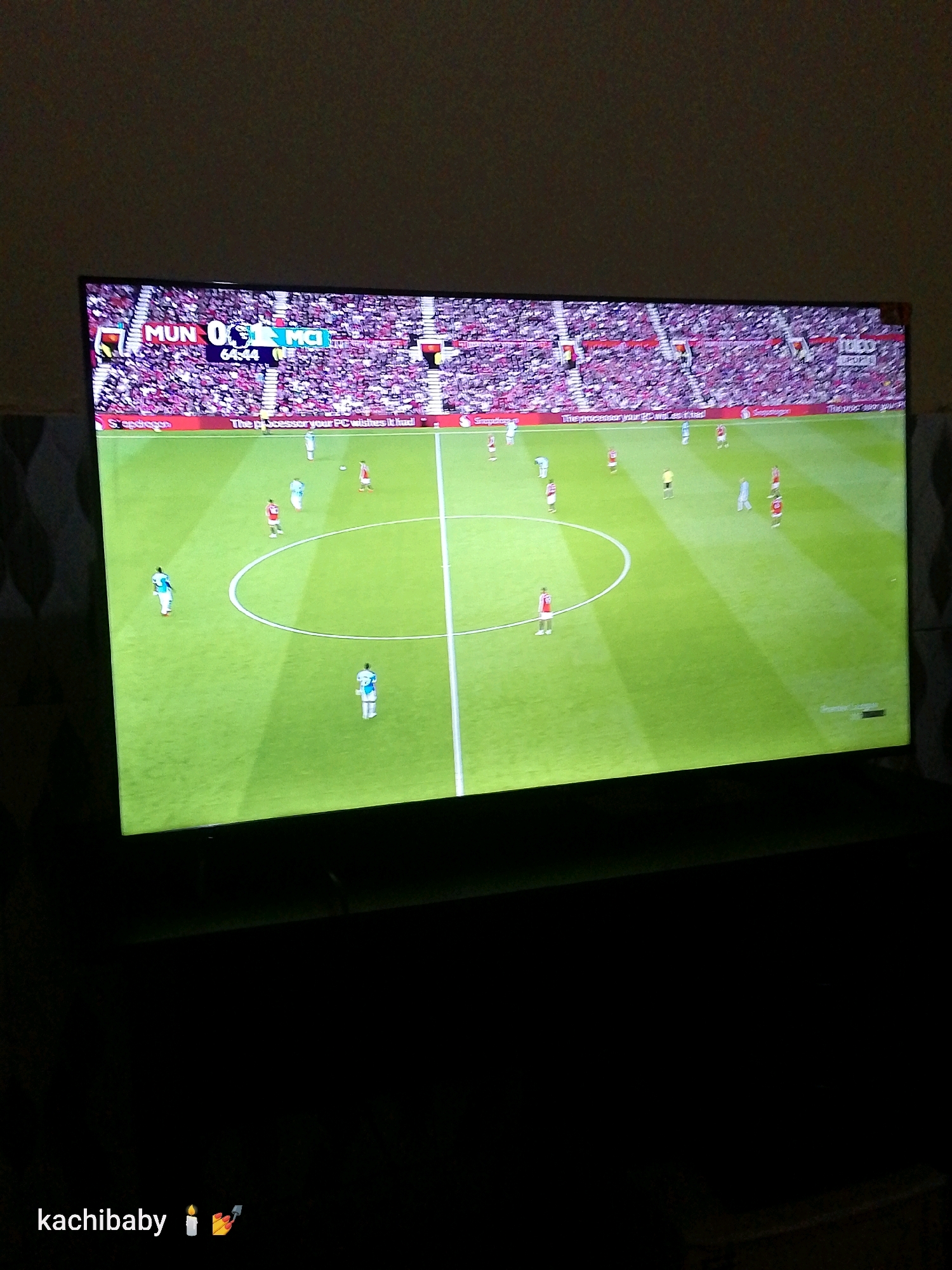 The image shows a television screen displaying a soccer match between two teams, identified by the abbreviations "MUN" and "MCI" on the scoreboard. The score is shown as 0-0, and the game time is 64:44. The stadium appears to be full of spectators wearing red and white attire. The field is green, with white lines marking the boundaries and center circle. Several players are visible on the field. In the foreground, the text "kachibaby" is overlaid with emojis of a candle and a celebration popper, suggesting a personal context. The background is dark, indicating a dimly lit room or nighttime viewing. There is no indication of the specific location within Mtopanga ward, Kenya, or any other geographic details relevant to that ward.