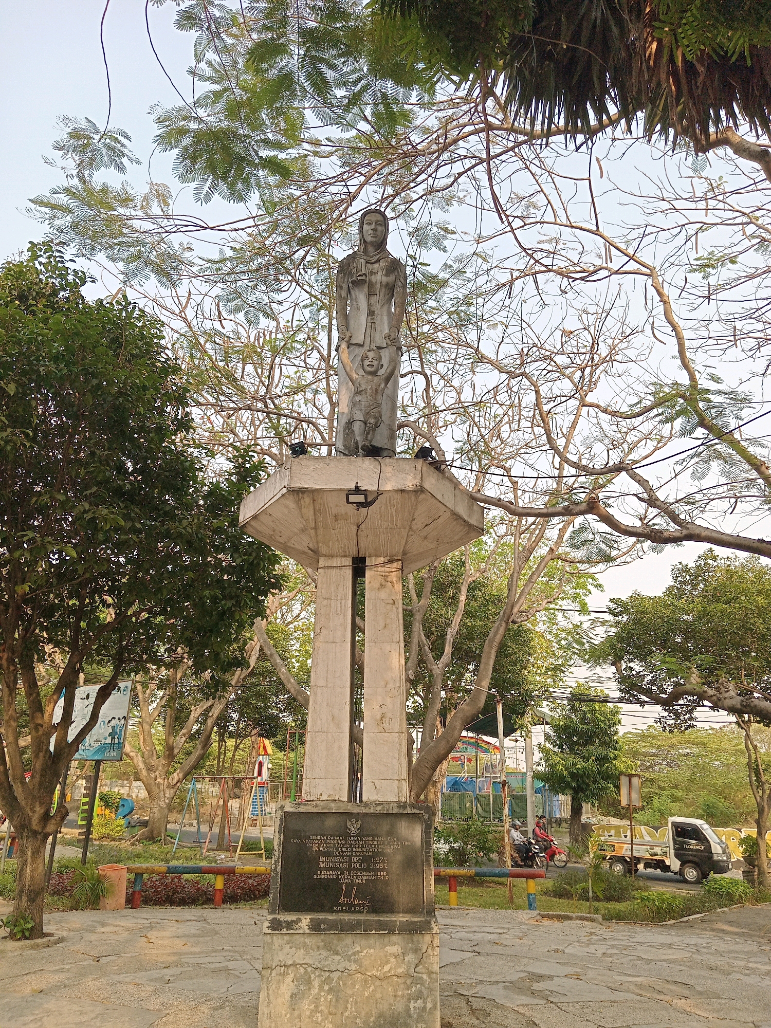 A statue of a woman holding a child stands atop a concrete pedestal in what appears to be a park in Randuagung, Indonesia. The statue is made of concrete and depicts the woman with a head covering and the child with arms raised. The pedestal has a plaque at its base with text in Indonesian, indicating it is a monument. Surrounding the statue, there are trees with sparse foliage, a playground with swings and a slide, and a paved area. In the background, there is a white and black truck, a motorcycle with two riders, and other vegetation. The sky is overcast. The scene suggests a public space with recreational facilities.