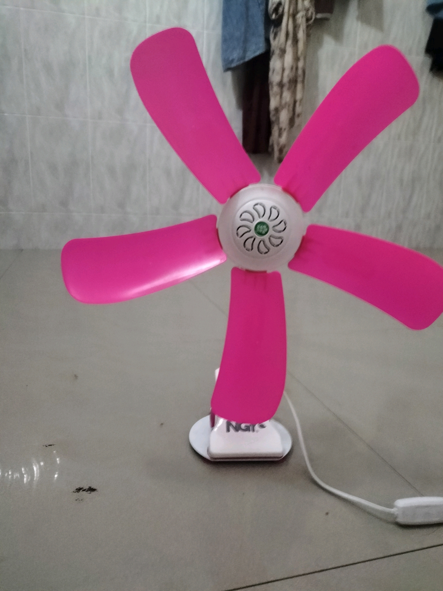 A pink electric fan with five blades is positioned on a light gray floor. The fan has a white circular motor housing with decorative grilles and a green logo in the center. The fan's base is white and rectangular, with the letters "NGY" visible on its side. A white power cord extends from the base of the fan to a white in-line switch. The background consists of a tiled wall with a grid pattern. Hanging on the wall are various pieces of fabric, possibly clothing. The image appears to be taken indoors, in what could be a domestic setting. There are no people visible in the image. The lighting is somewhat dim, casting faint shadows on the floor.