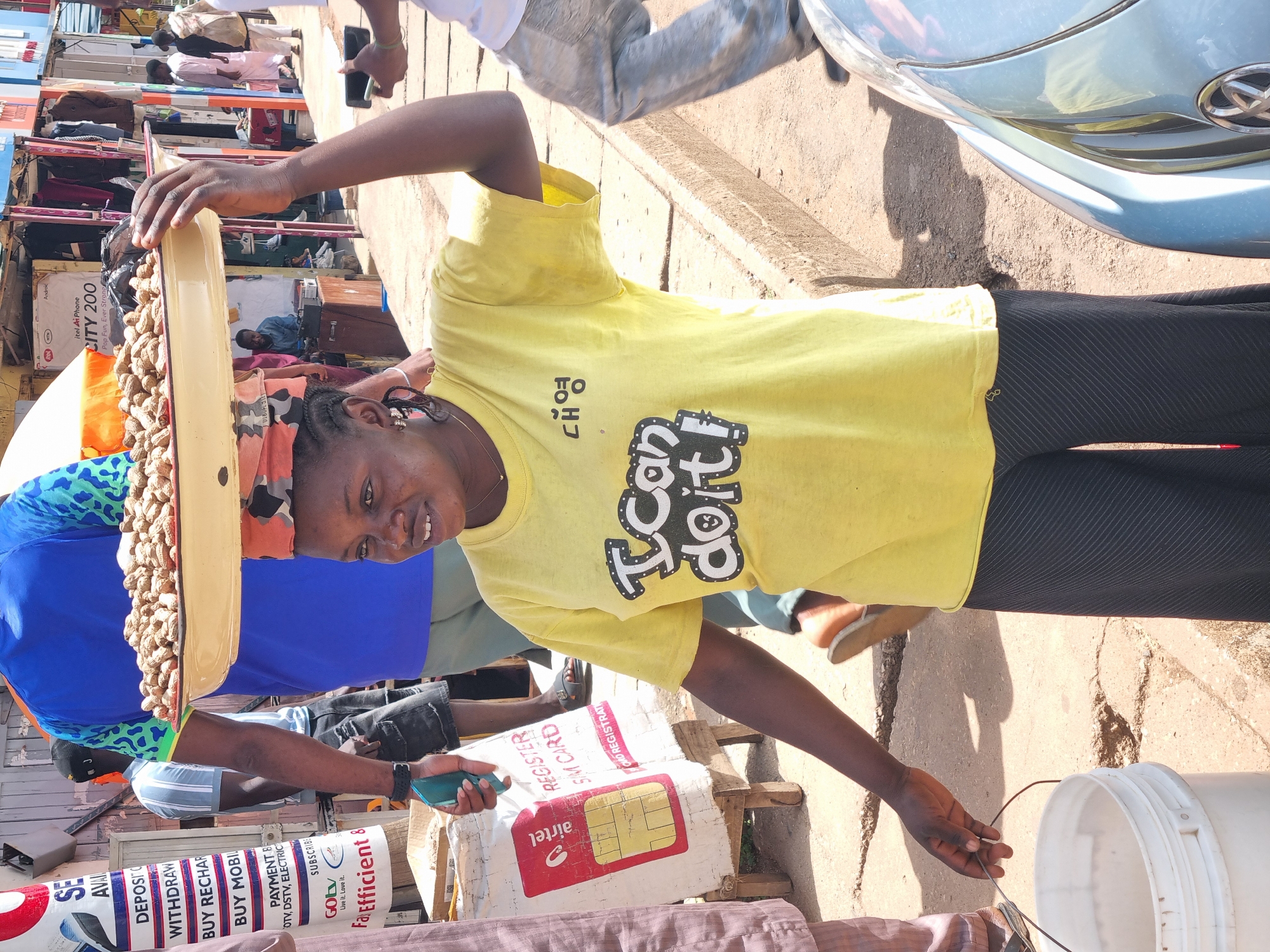 The image depicts a street scene in Jos, Nigeria, on a sunny day. A young woman, wearing a bright yellow t-shirt with a graphic print and dark, striped pants, balances a large, shallow tray of peanuts on her head. She is holding a white bucket with her left hand. To her left, another person is partially visible, wearing a brown garment and holding a bag with the Airtel logo. Behind her, a man in a blue shirt and another in a blue shirt with a patterned collar are visible, along with a display of various goods for sale. The environment appears to be a bustling market or street-side stall area, with paved ground and a concrete curb. In the foreground, the fender and headlight of a light blue car are visible.
