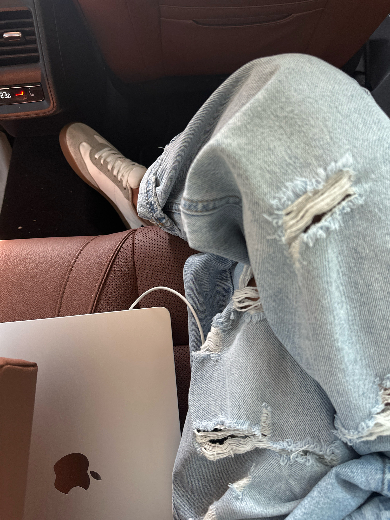 The image depicts a close-up view of a person's legs wearing ripped light blue jeans, sitting in what appears to be the interior of a car. A silver laptop with an Apple logo is resting on a brown leather seat in the foreground. To the left, a portion of a car's climate control panel with a red digital display showing "23.0" is visible. The interior of the car has brown leather seats and dark carpeting. The lighting suggests an indoor setting, likely during the daytime. There is no information within the image to determine the specific location within the city of Sofia, Bulgaria.