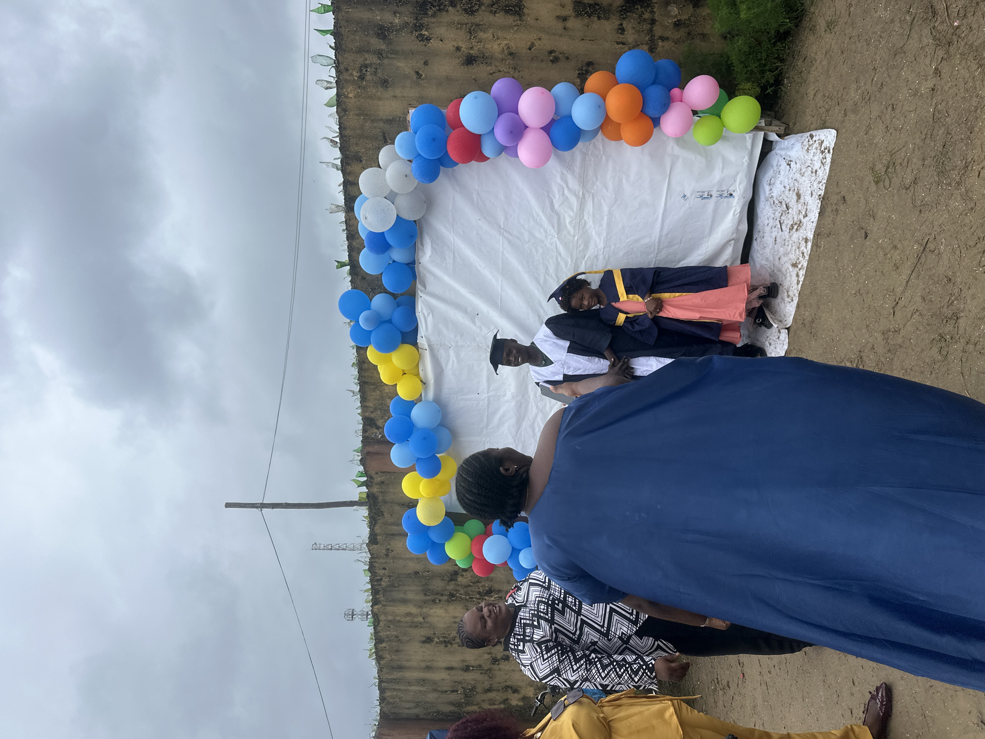 The image depicts an outdoor graduation celebration in Badagry, Nigeria. A balloon arch, comprised of blue, yellow, red, purple, pink, green, and orange balloons, frames a white backdrop. Two individuals in graduation attire, a young man and a young girl, stand side-by-side in front of the backdrop. The young man wears a black and white graduation gown with a cap, while the young girl wears a blue graduation gown with a yellow sash and a cap.
Several adults are present, some facing the graduates and others turned away from the camera. To the left, a woman in a black and white patterned top and a woman in a large blue dress are visible. Another woman in a yellow outfit is partially visible on the far left. The background features a weathered concrete wall topped with broken glass shards, a common security measure in some areas. Power lines and a distant tower can be seen against a cloudy sky. The ground appears to be dirt or sand, with some scattered debris. The scene suggests a festive but informal gathering, likely in a residential or community area in Badagry.